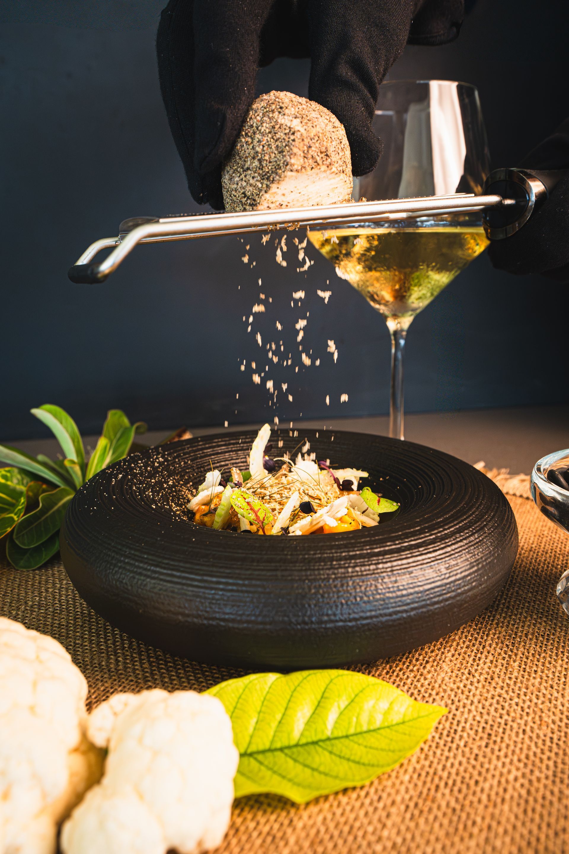 A chef grating seasoning over a plate of food, with a glass of wine in the background.