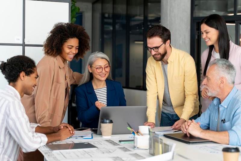 Team of diverse people collaborating around a laptop in an office, smiling and looking engaged.