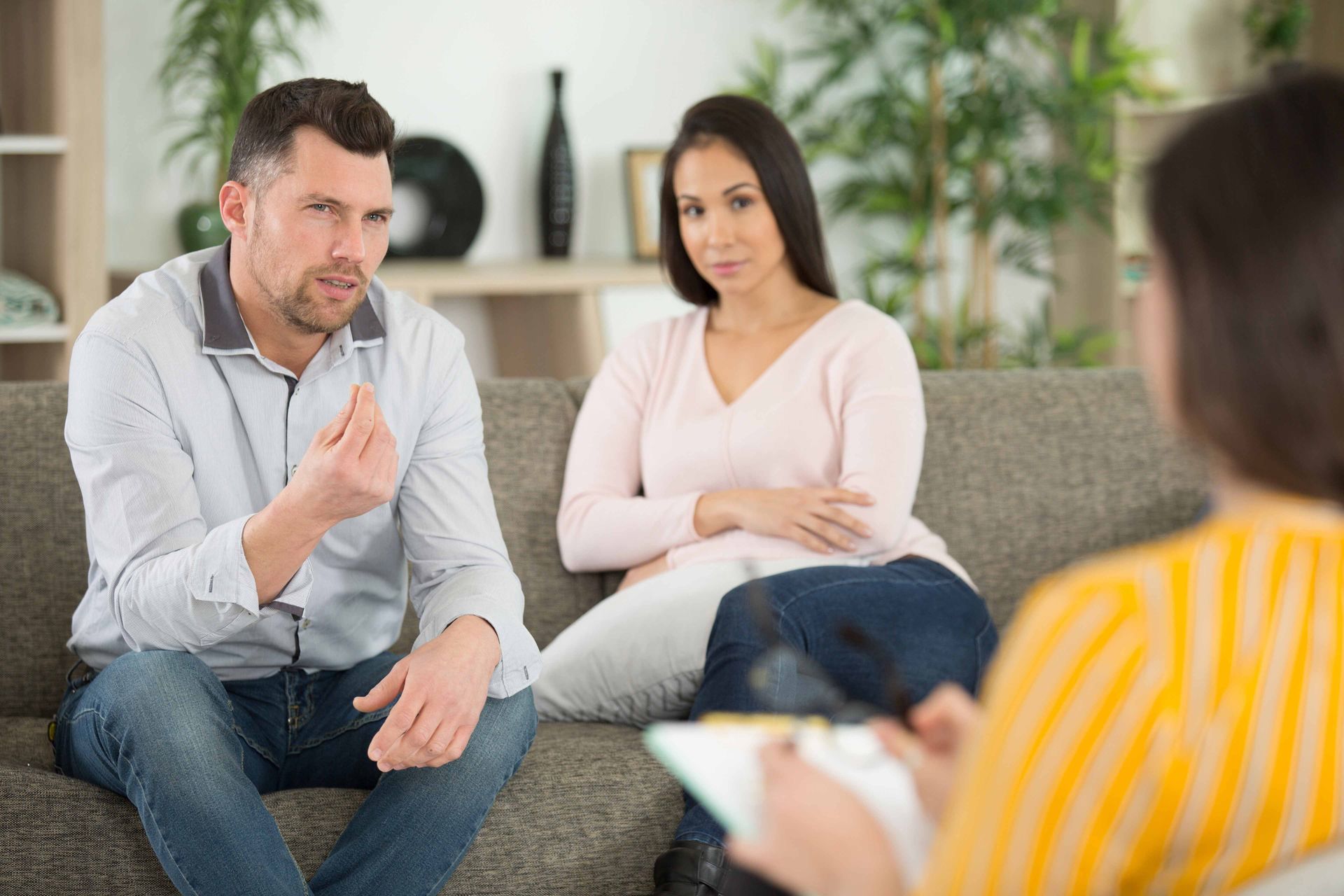 Couple in therapy session, man gesturing, woman looking on, therapist taking notes.