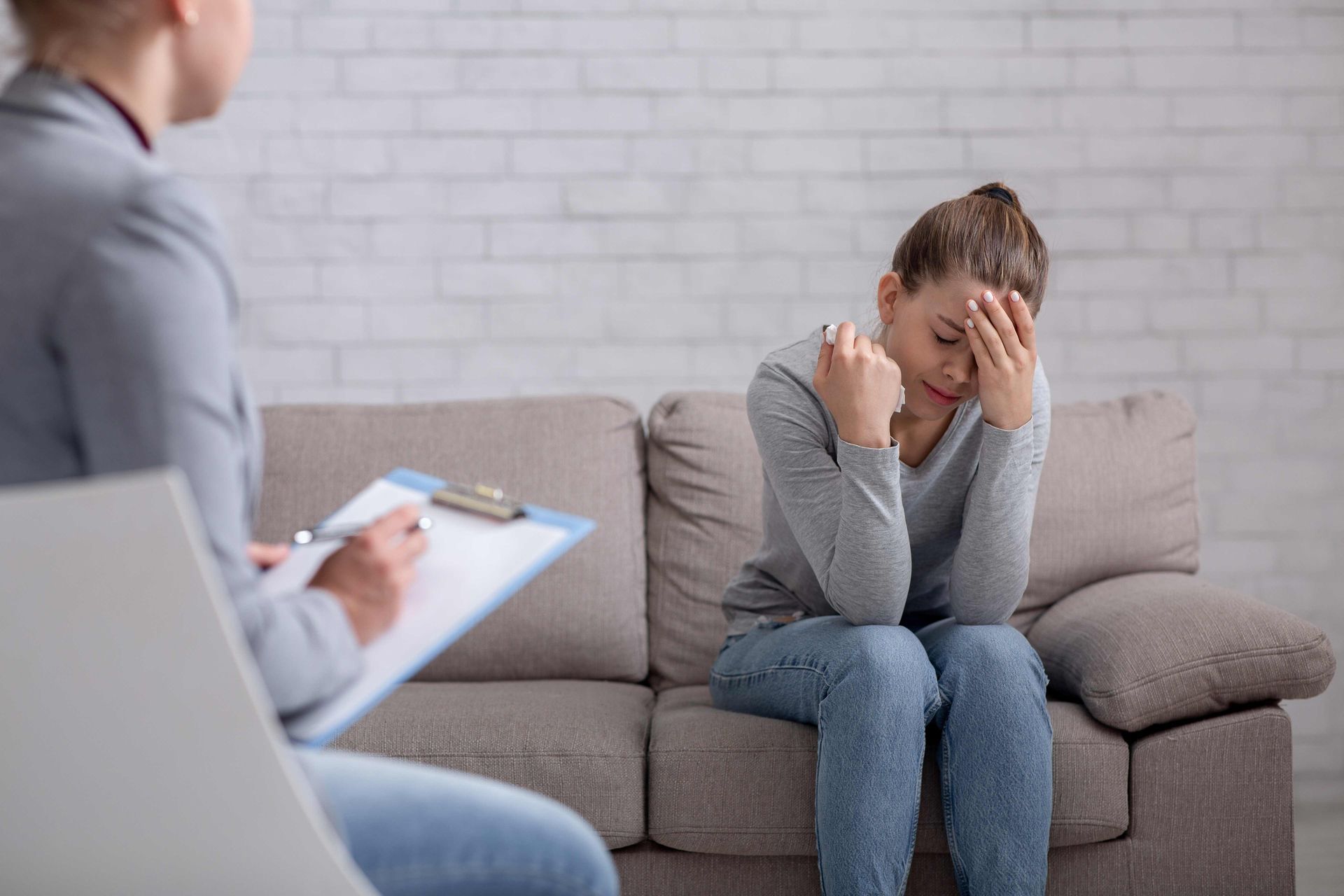 Woman on couch with head in hands during therapy session. Therapist taking notes.
