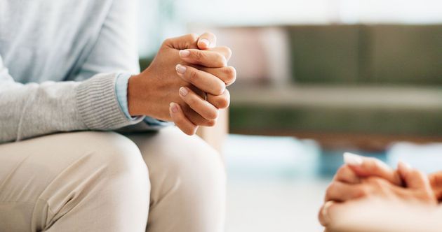 Person with hands clasped, seated in a counseling session; a second person's hands are visible. Person with hands clasped, seated in a counseling session; a second person's hands are visible.