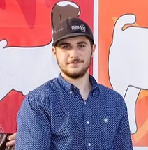Young man wearing a baseball cap and button-down shirt, smiling, standing outdoors near a colorful dog illustration.