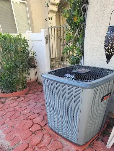 AC unit on red brick patio next to a white fence and greenery.