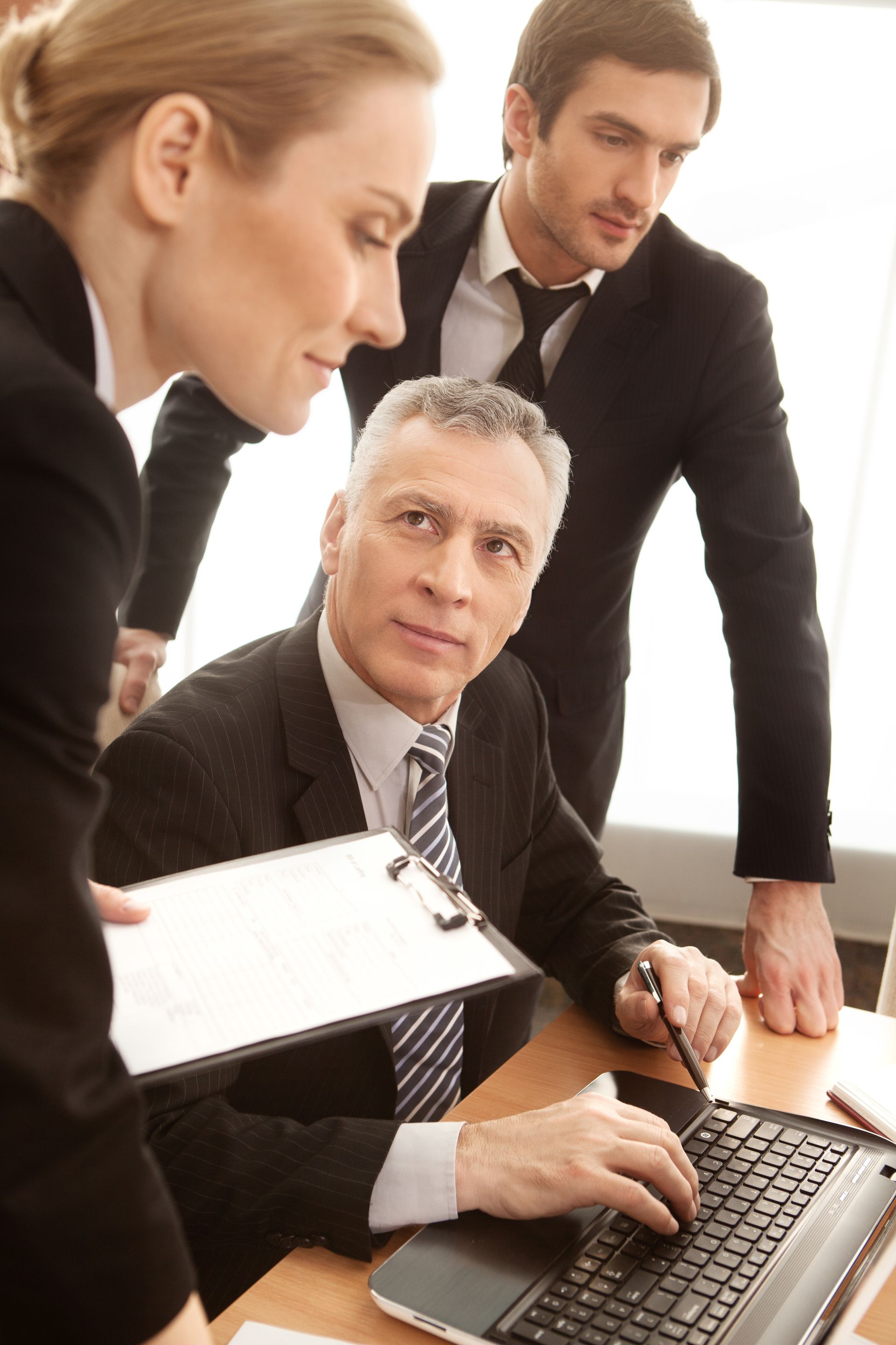 Three business colleagues gathered around a laptop in an office, reviewing documents and discussing work.