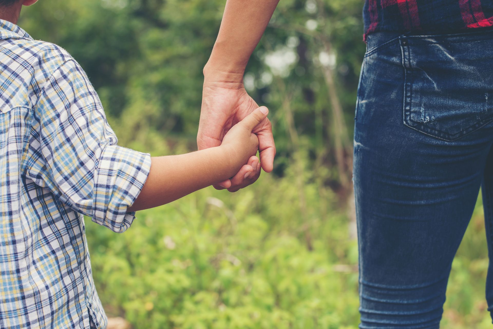 Adult holding a child's hand on a wooded path, both wearing plaid shirts and jeans