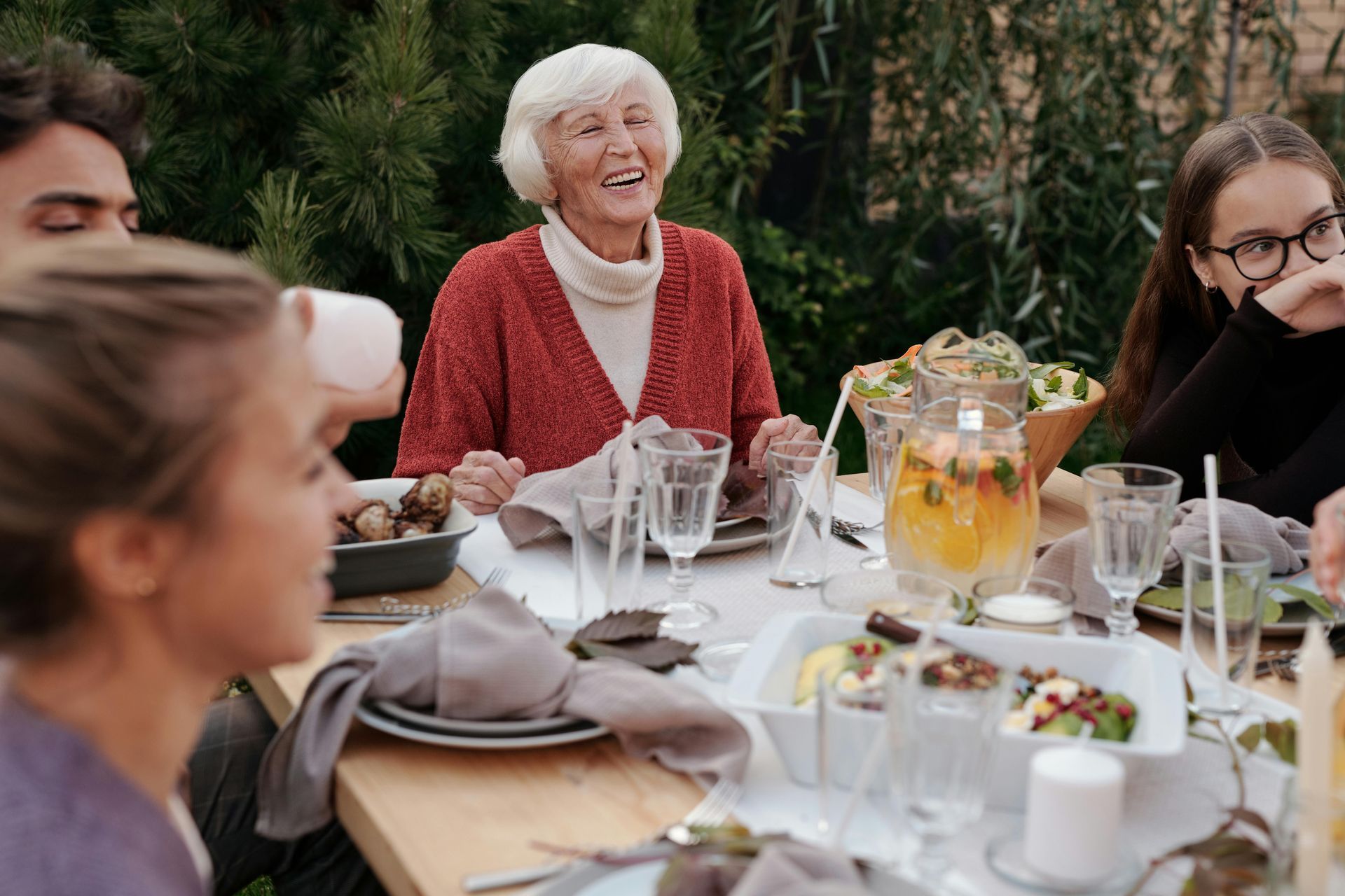 Family seated at outdoor table, elderly woman smiling, various dishes and drinks present.