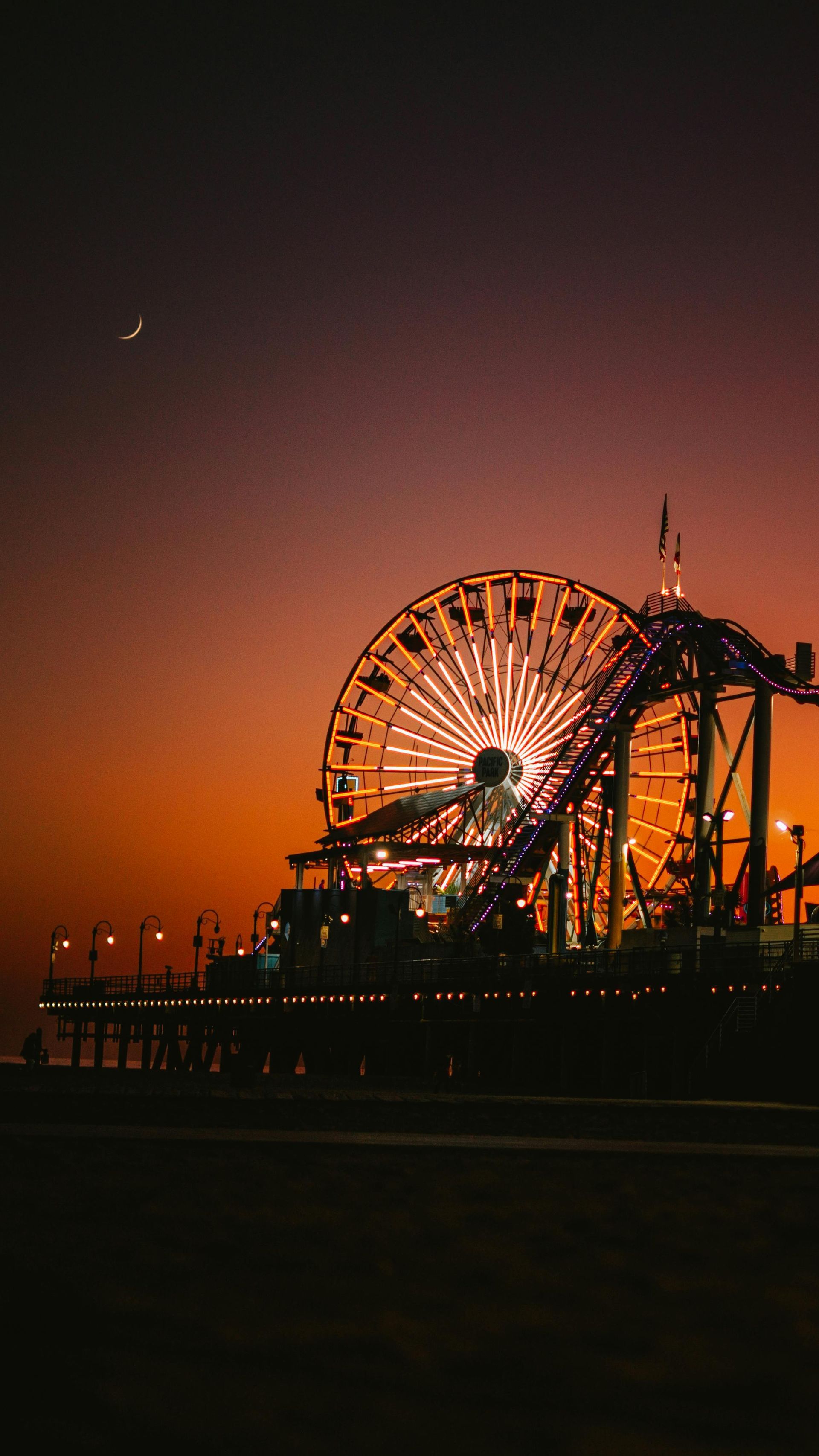 Ferris wheel and rollercoaster lit up at sunset on a pier, with orange and purple sky.