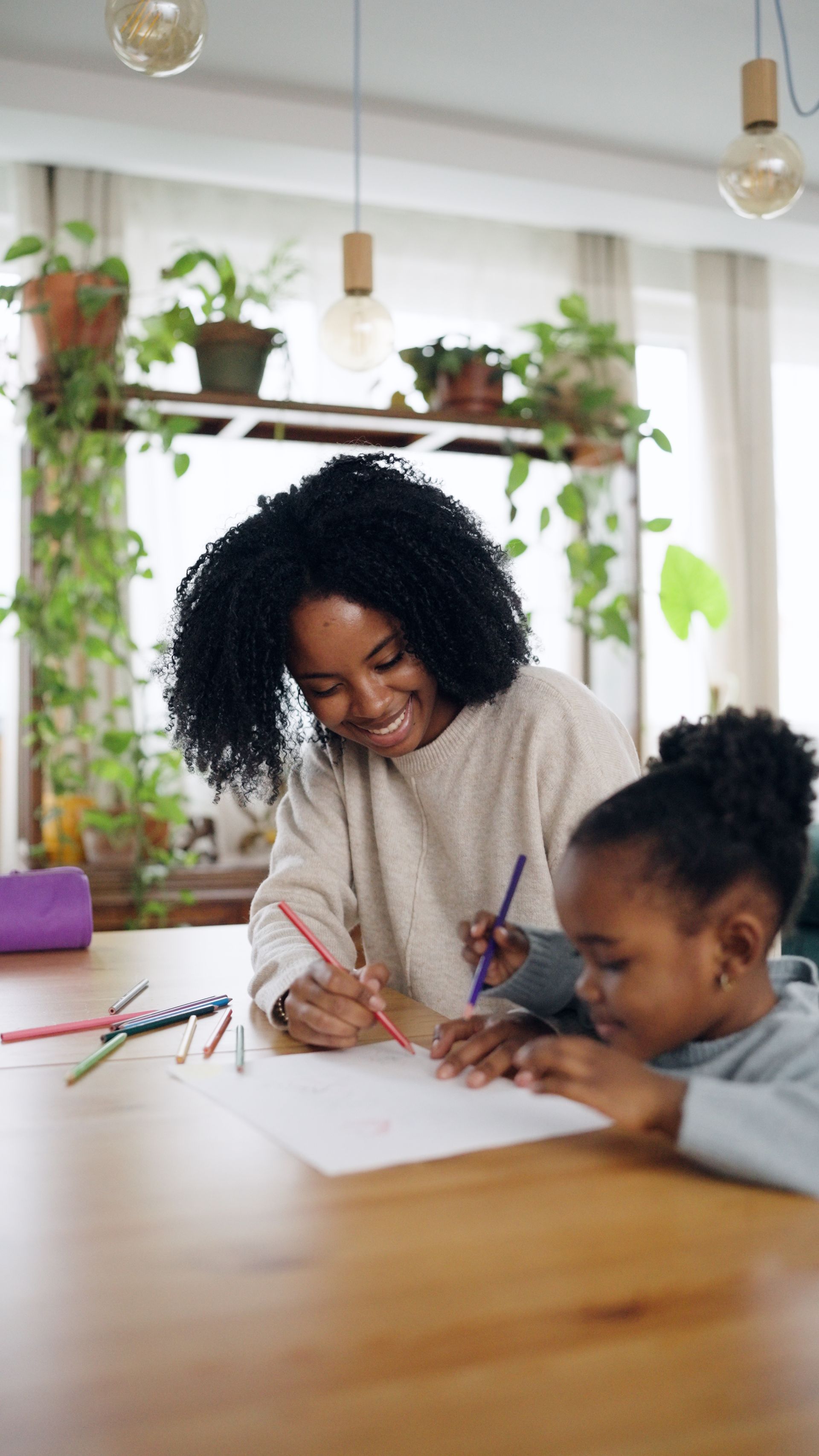 Adult and child drawing together at a wooden table in a bright home with plants and hanging lights