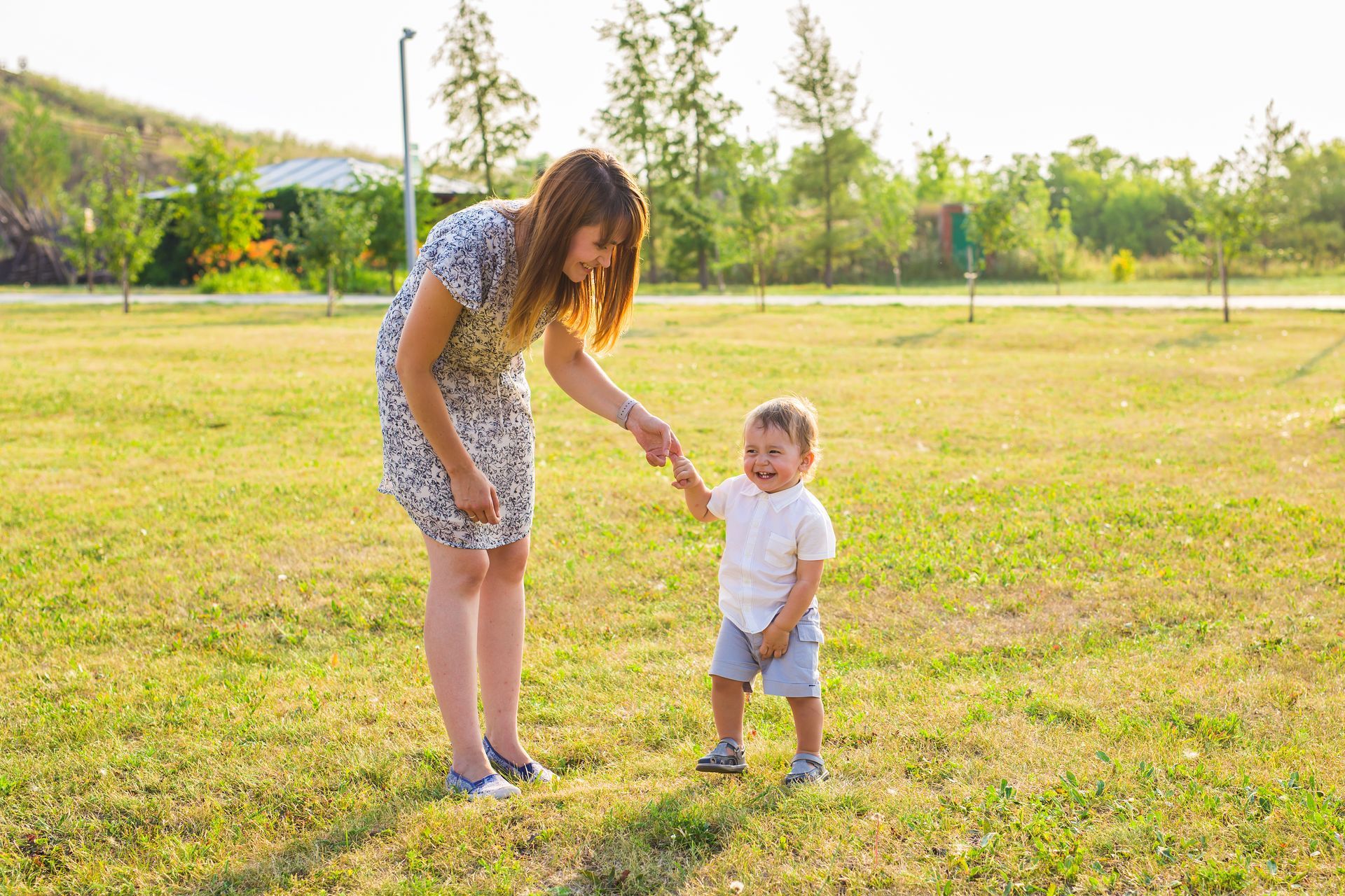 Adult helping toddler walk in a sunny park, both standing on grass near trees and a path.