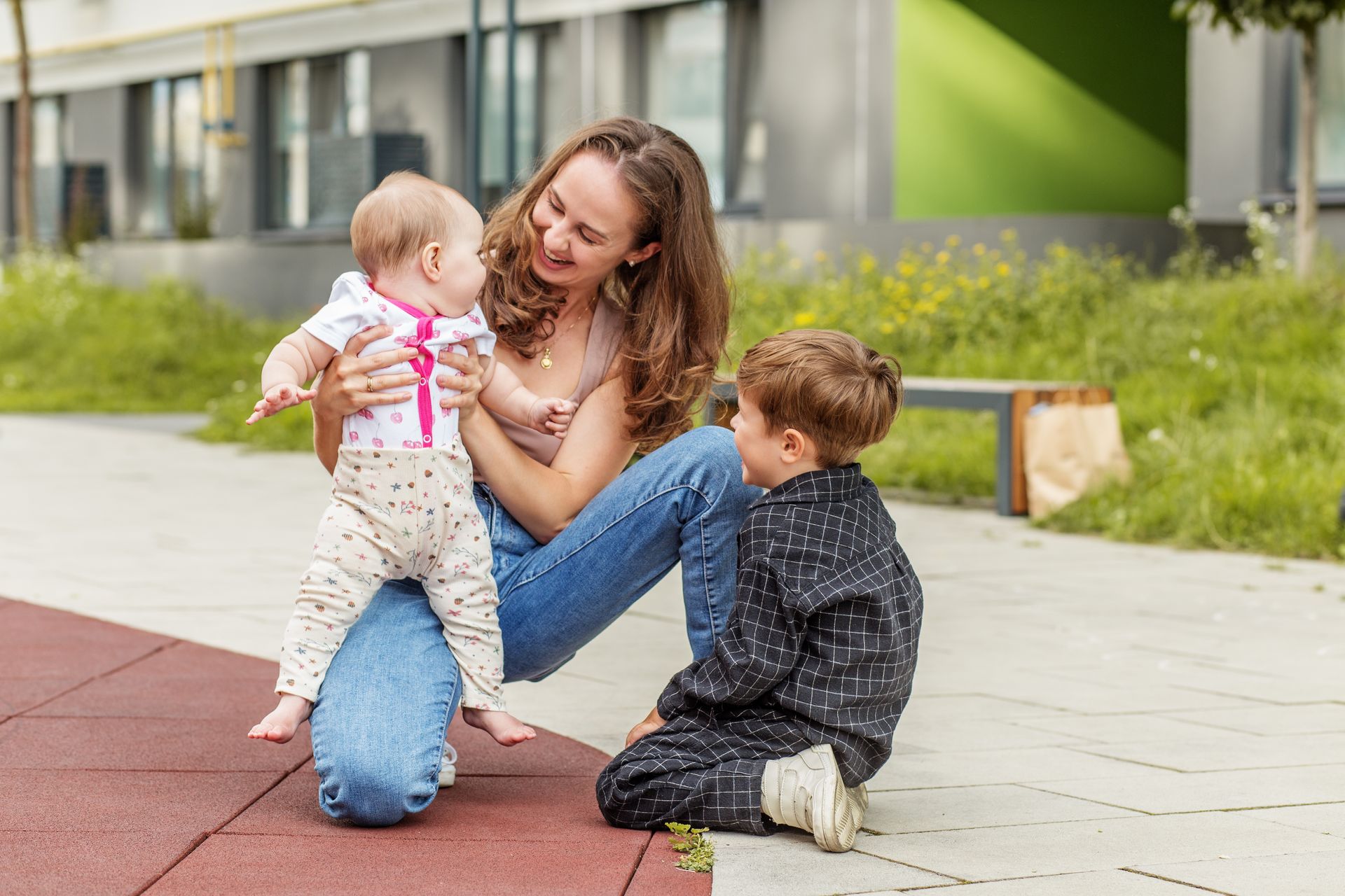 Woman kneeling on a path, smiling with two small children outdoors near a modern building.