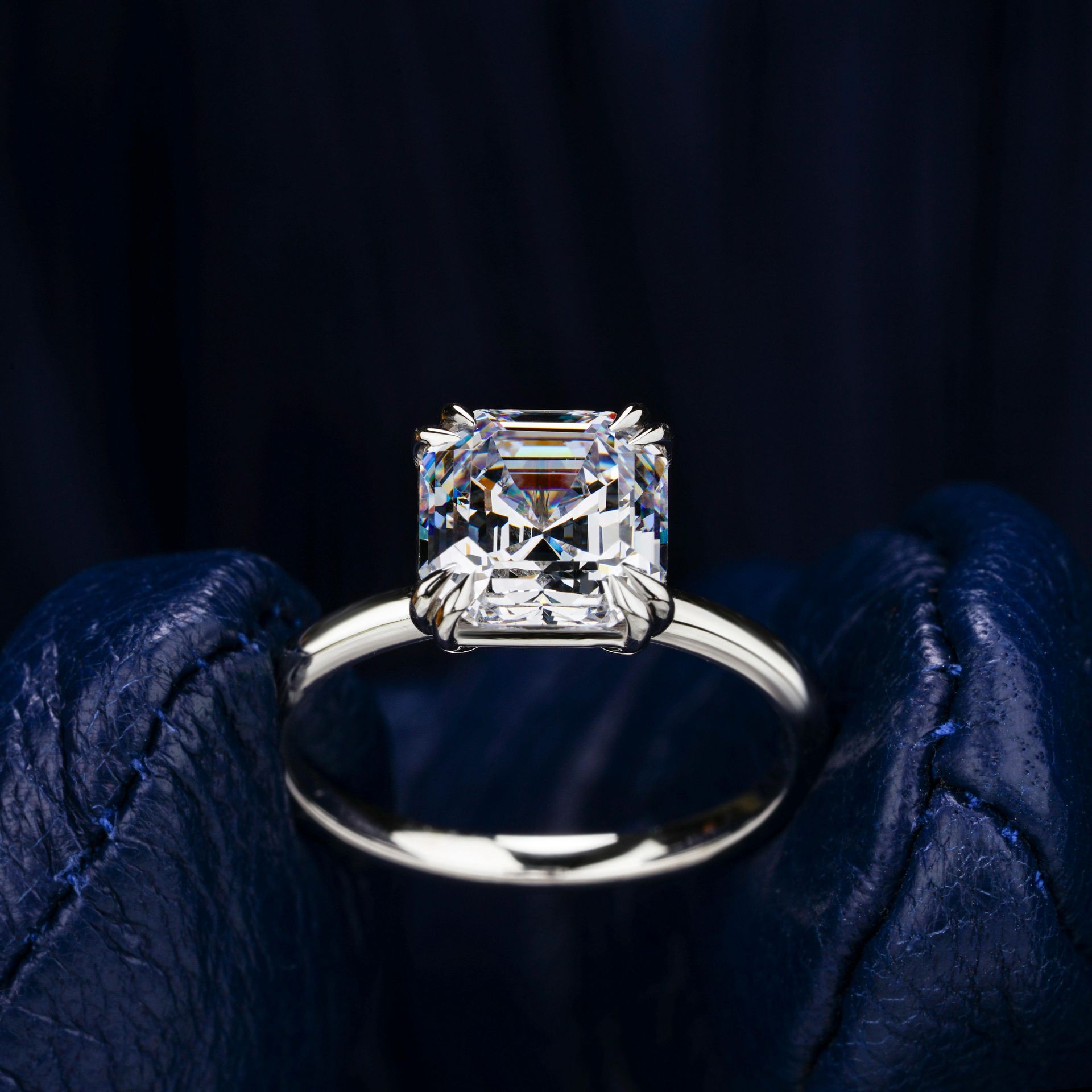 A brilliant-cut square diamond engagement ring held against a dark, textured blue background.