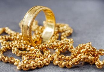 A textured gold ring resting on a pile of gold chain links against a grey stone background.