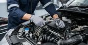 A mechanic wearing gloves uses a ratchet tool to work on a car engine in an auto repair shop.