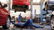 Two mechanics working in an auto repair shop; one lies on a creeper under a car while another stands at a workbench.