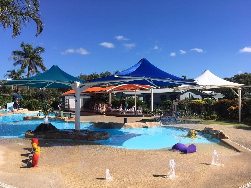 A Large Swimming Pool Surrounded by Umbrellas and Fountains on A Sunny Day — Jon Wilson Canvas & Shade in Coffs Harbour, NSW