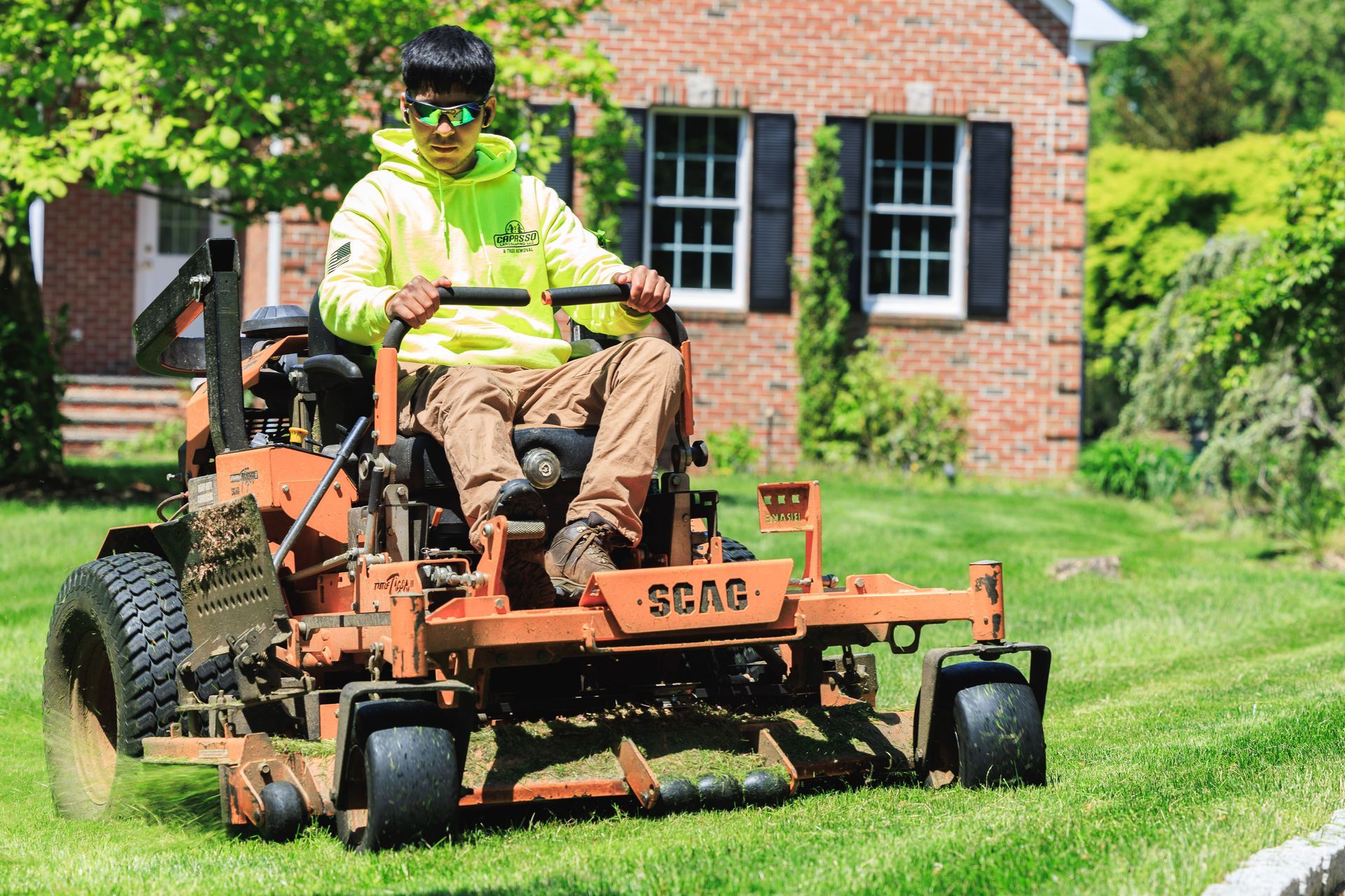 Person on a SCAG lawnmower mowing a green lawn in front of a brick house. They wear a safety vest and sunglasses.