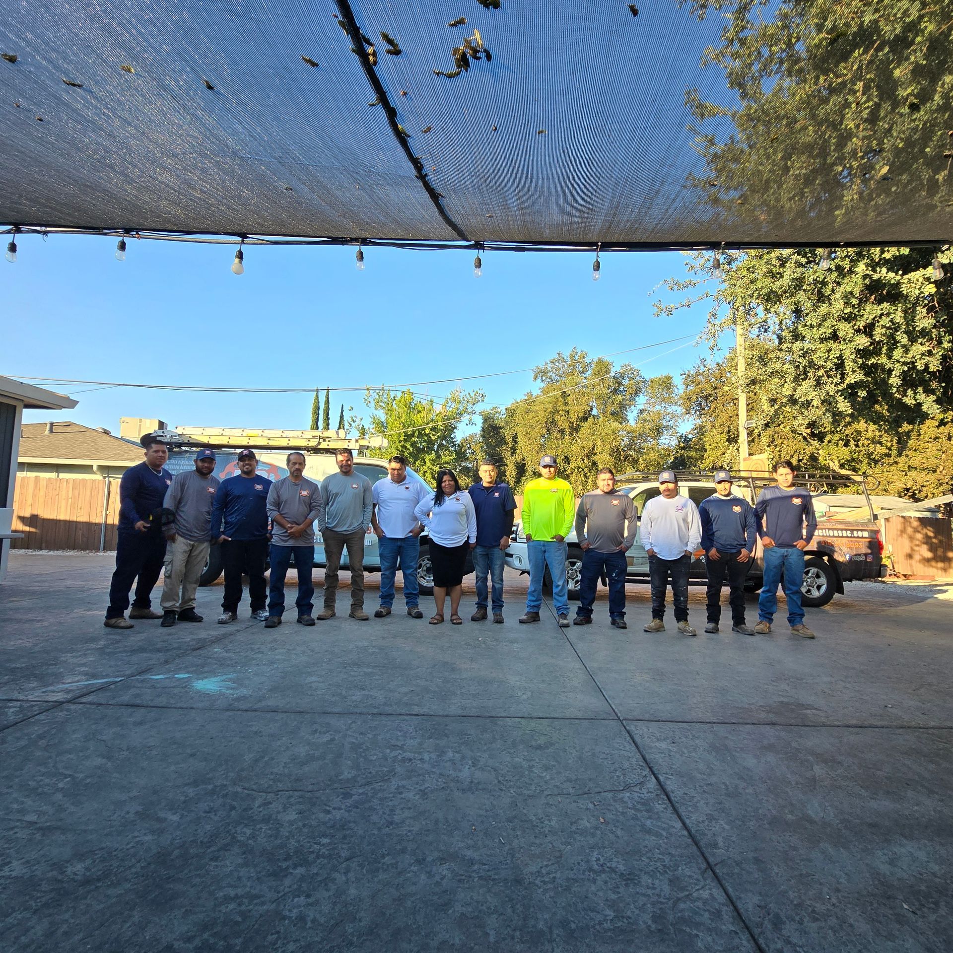 Group of workers standing in a yard under shade netting, with one person in a neon yellow shirt.
