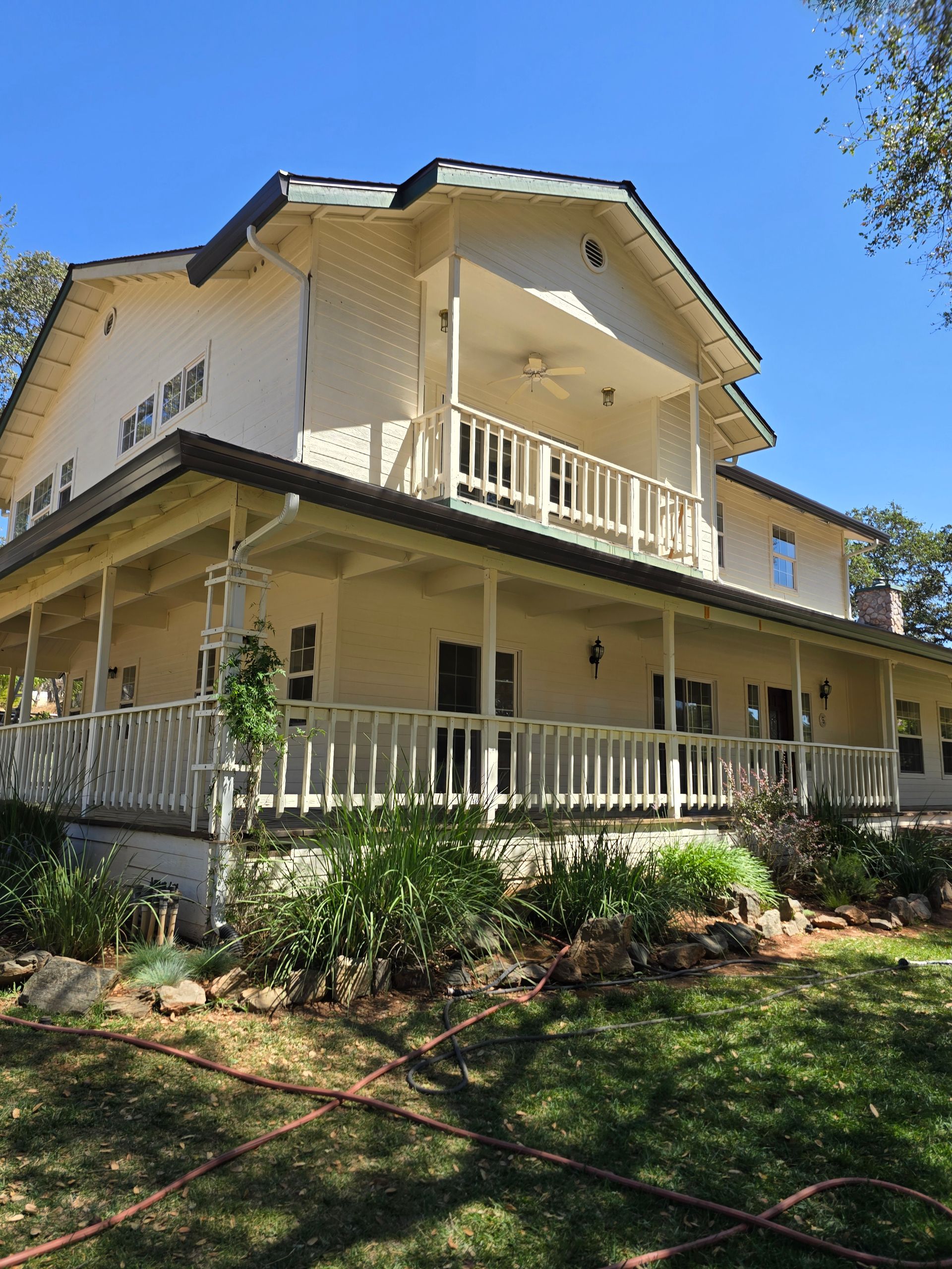 Two-story yellow house with wraparound porch, white railings, and garden on a sunny day