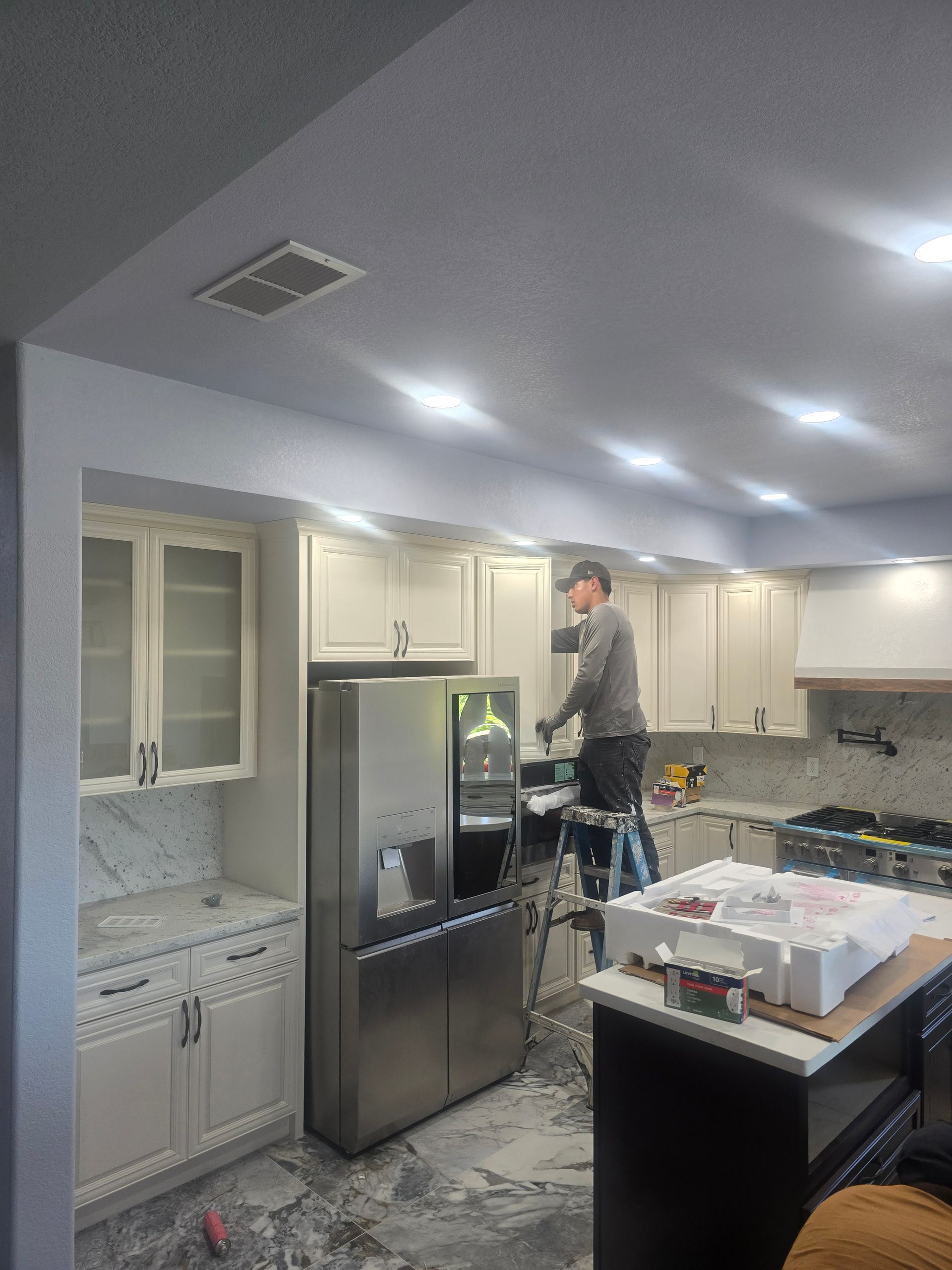 Person standing on a step stool adjusting cabinets in a bright kitchen with a stainless steel refrigerator.