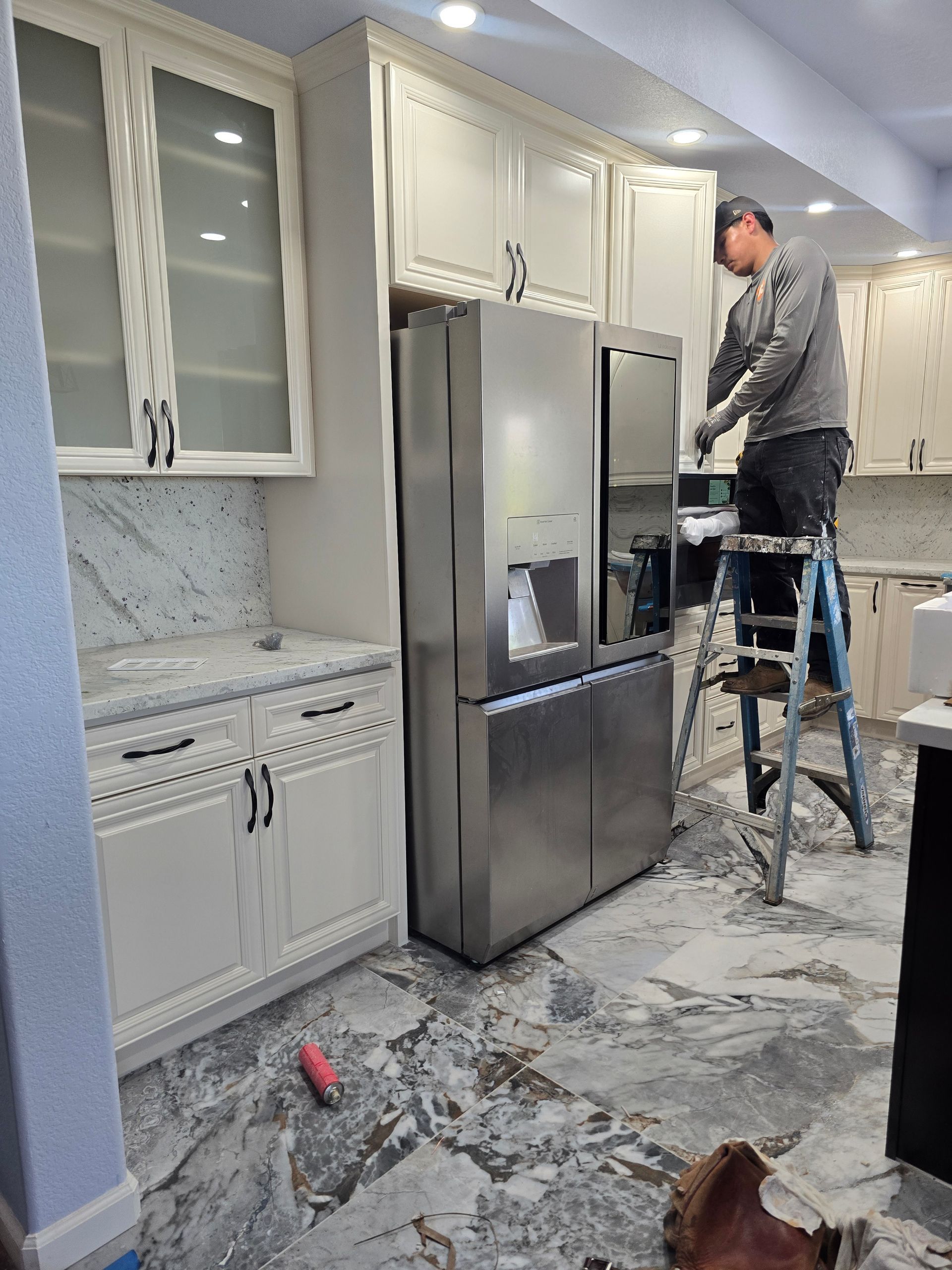 Worker on ladder installing cabinets in a bright kitchen with a stainless steel refrigerator.