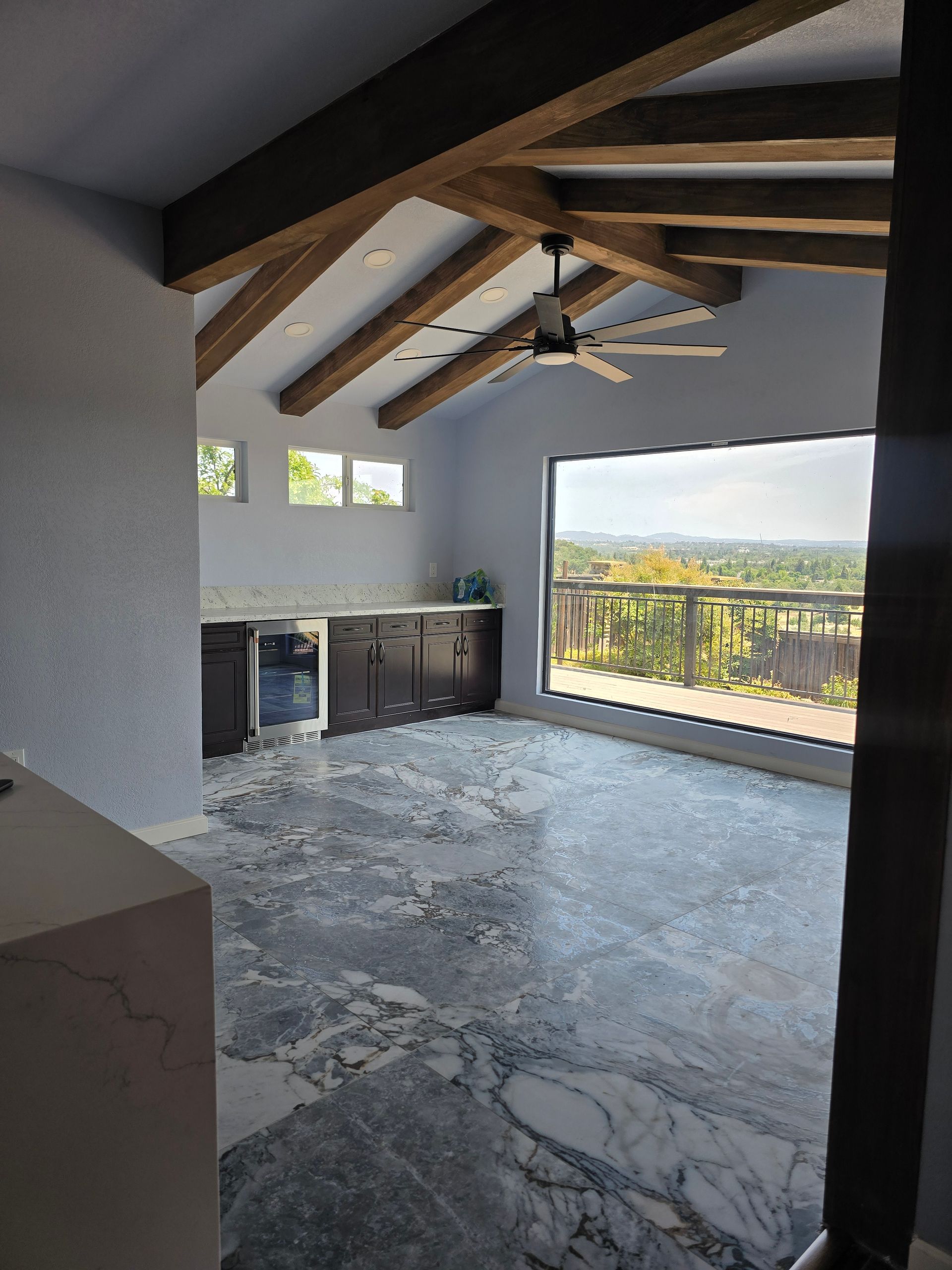 Empty room with exposed wooden beams, gray walls, and a large open view to a balcony and hills.