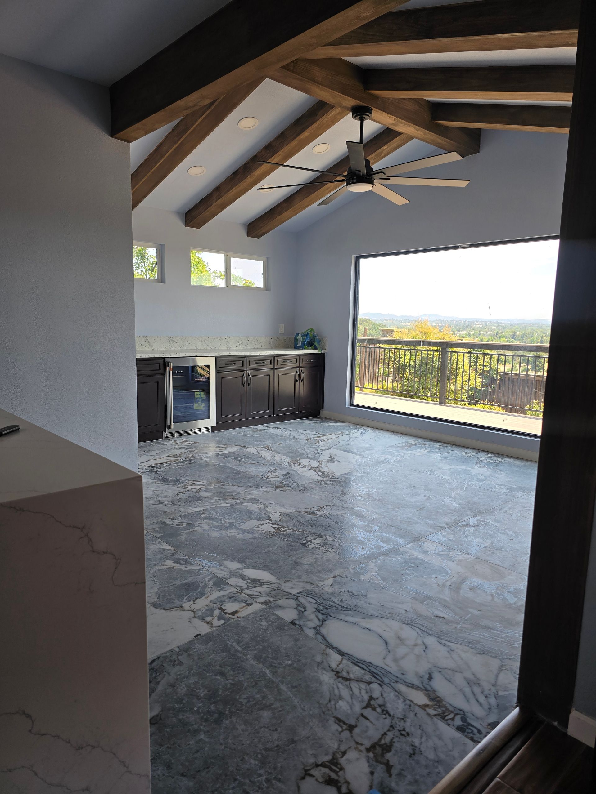 Modern room with gray stone floor, exposed wood beams, a ceiling fan, and a large view of the outdoors.