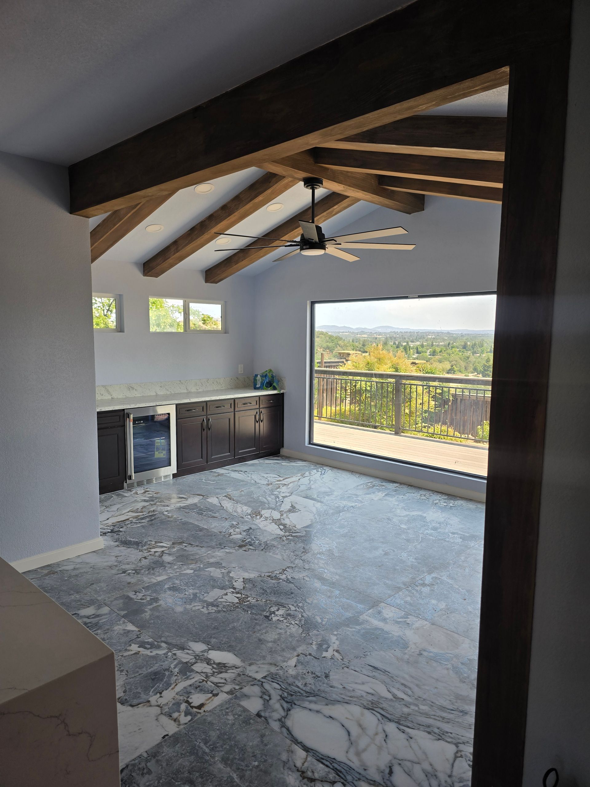 Empty modern room with exposed wooden ceiling beams, marble floor, and large windows overlooking a balcony.