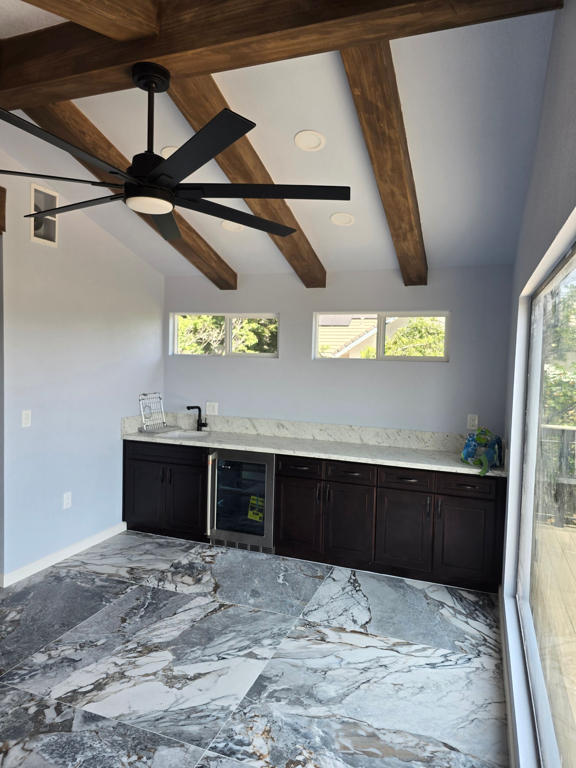 Modern kitchen with dark cabinets, stone countertop, large ceiling fan, and exposed wood beams.
