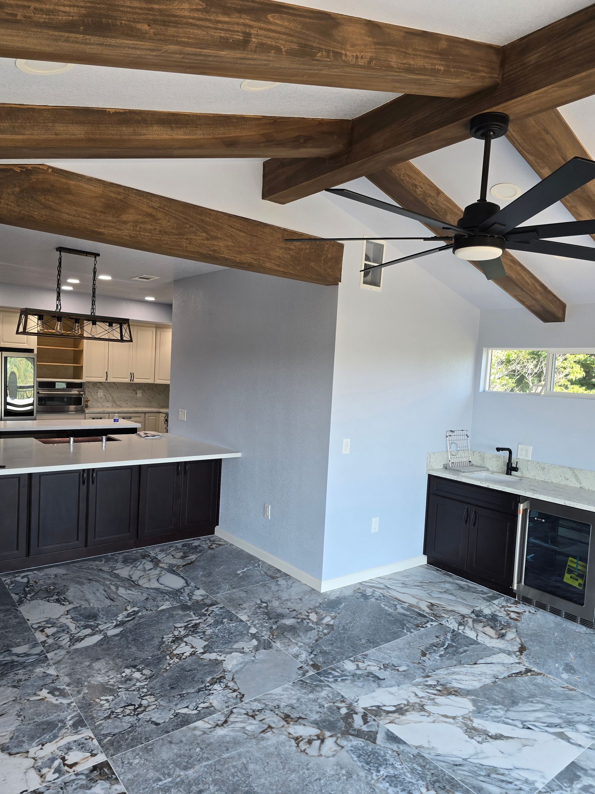 Open kitchen and living area with exposed wood beams, ceiling fan, dark cabinets, and light gray floor.