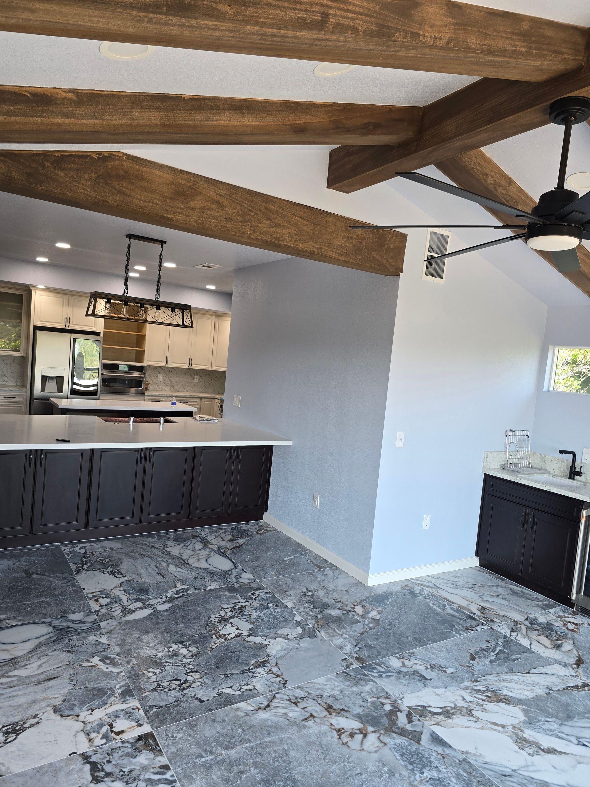 Open-plan kitchen with white walls, dark cabinets, stone floor, and exposed wood ceiling beams