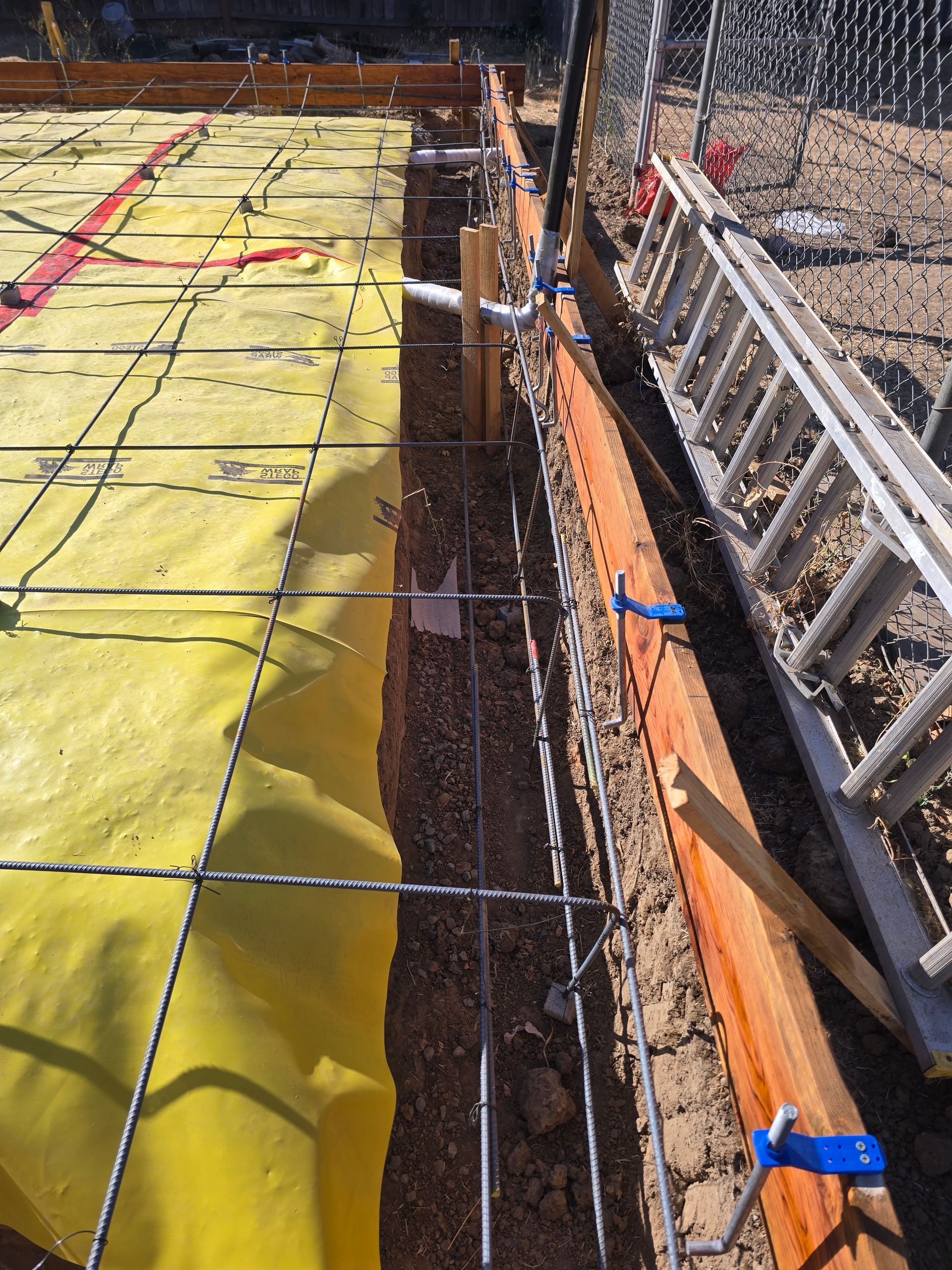 Construction site with rebar, orange formwork, and yellow waterproofing along a trench edge
