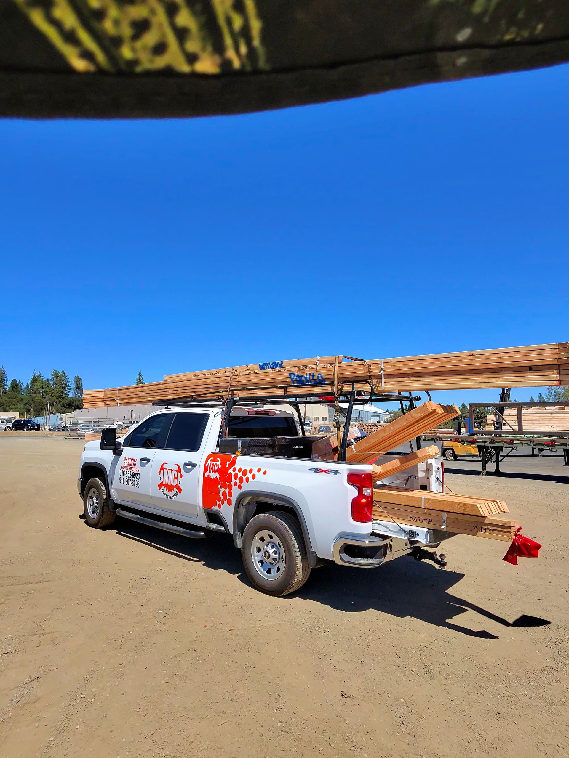 White pickup truck with red markings hauling a wooden ladder on a sandy lot under a bright blue sky