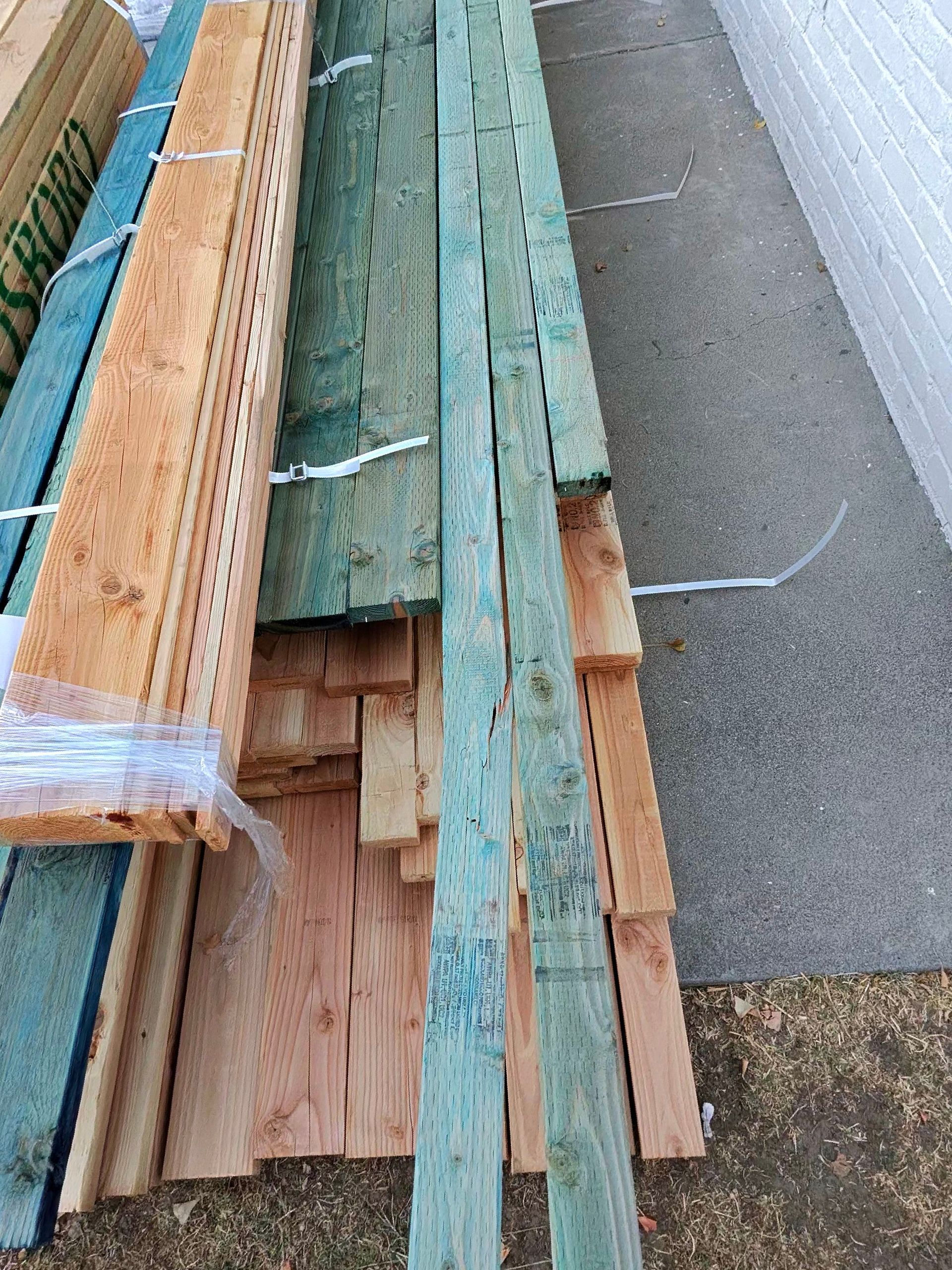 Stacked reclaimed wood planks beside a gray wall and green wire fence outdoors.
