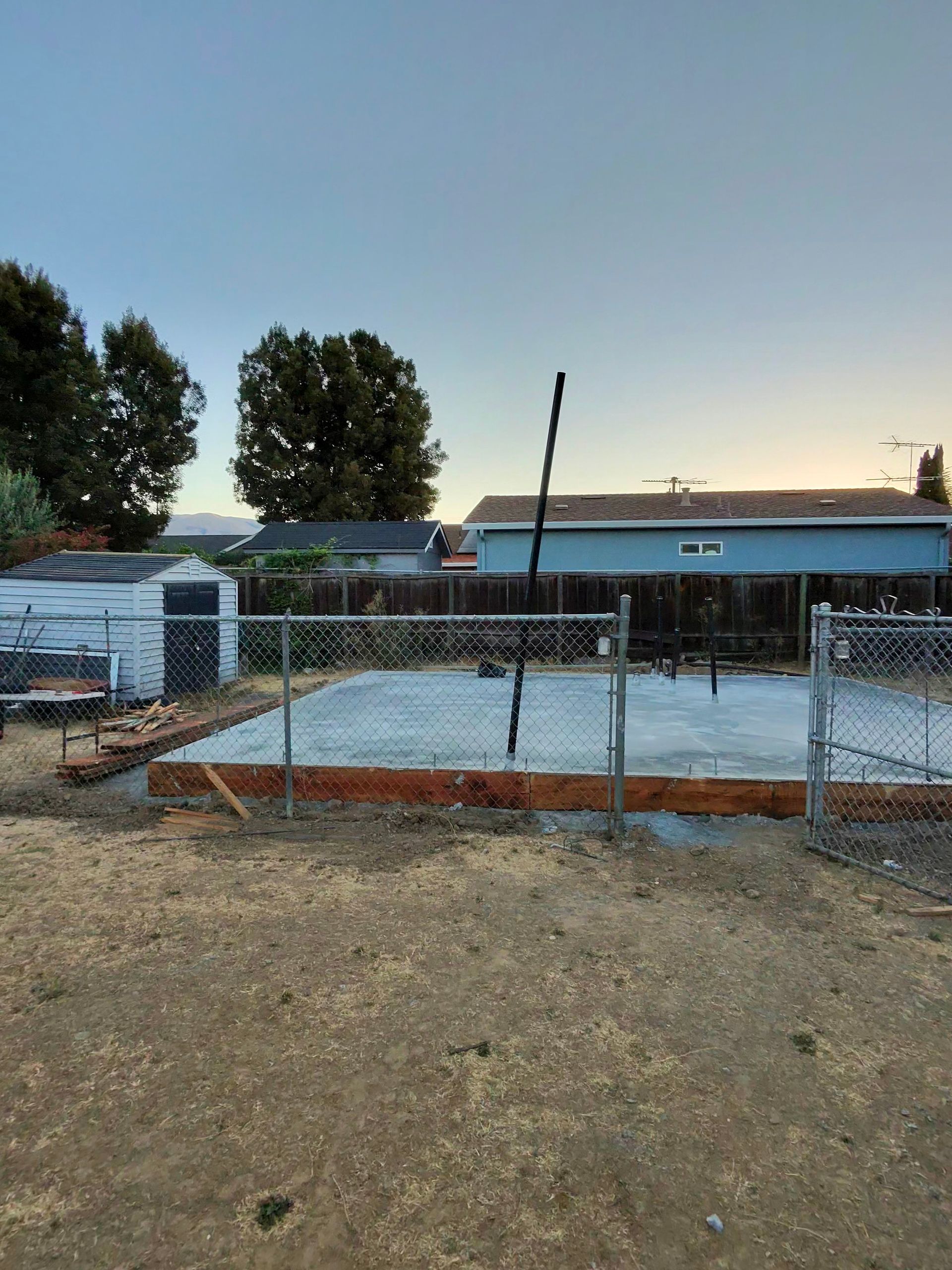 Chain-link fenced dirt lot with a concrete slab and low buildings at dusk, trees in the background