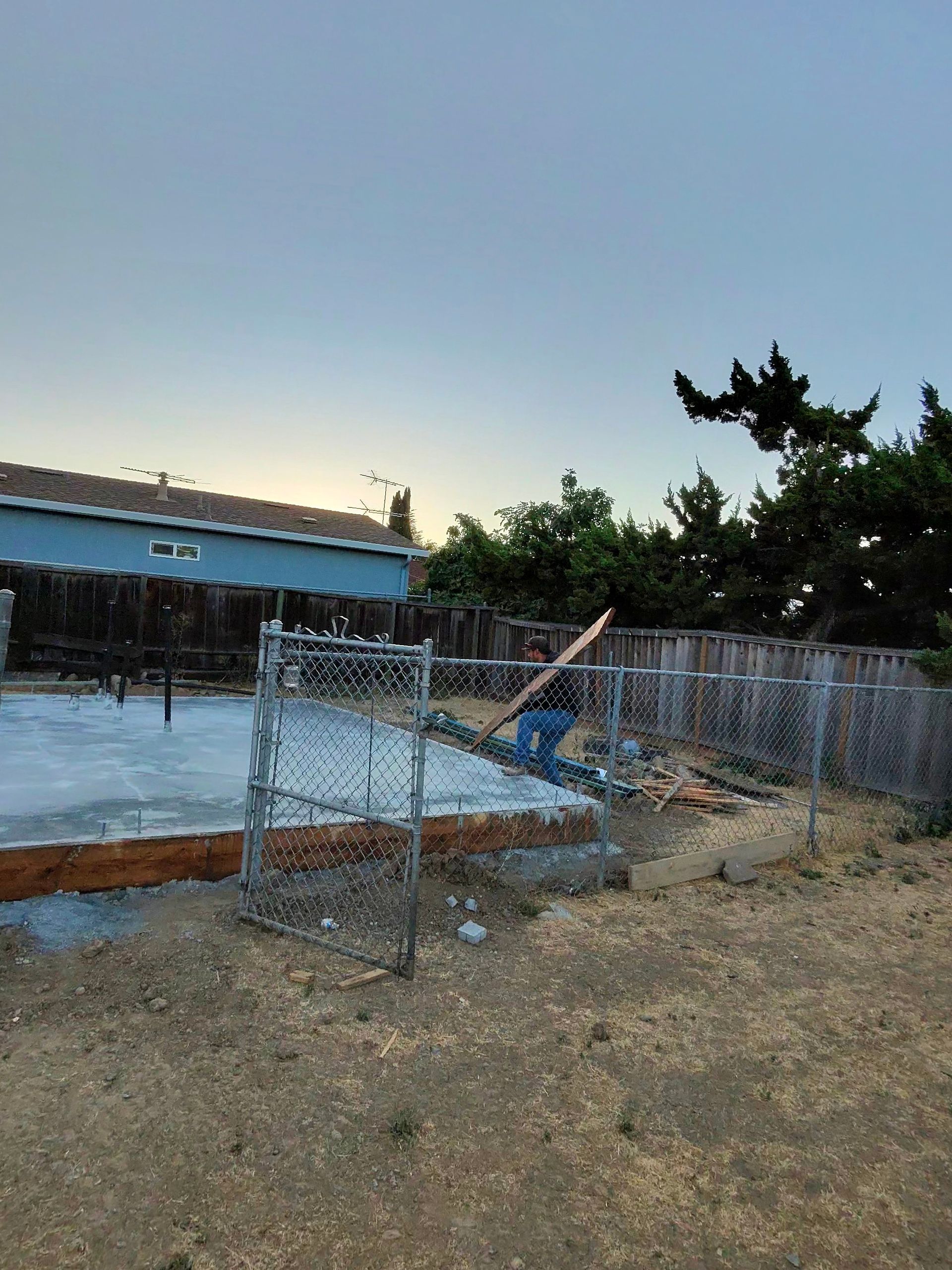 Chain-link fenced lot with a long blue-roofed building at dusk and a muddy foreground