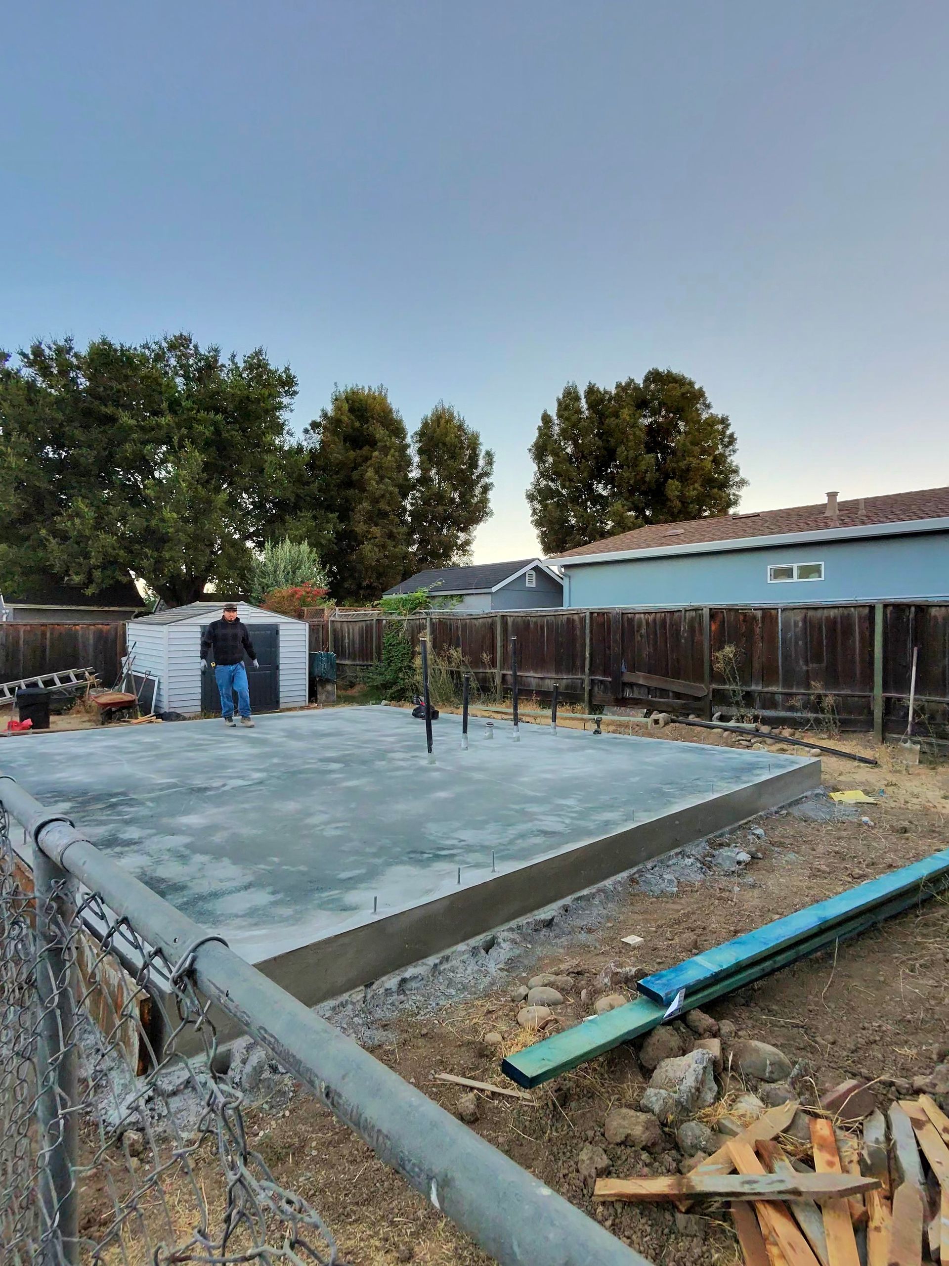 Concrete foundation being poured at a construction site, with workers, trees, and a small building nearby