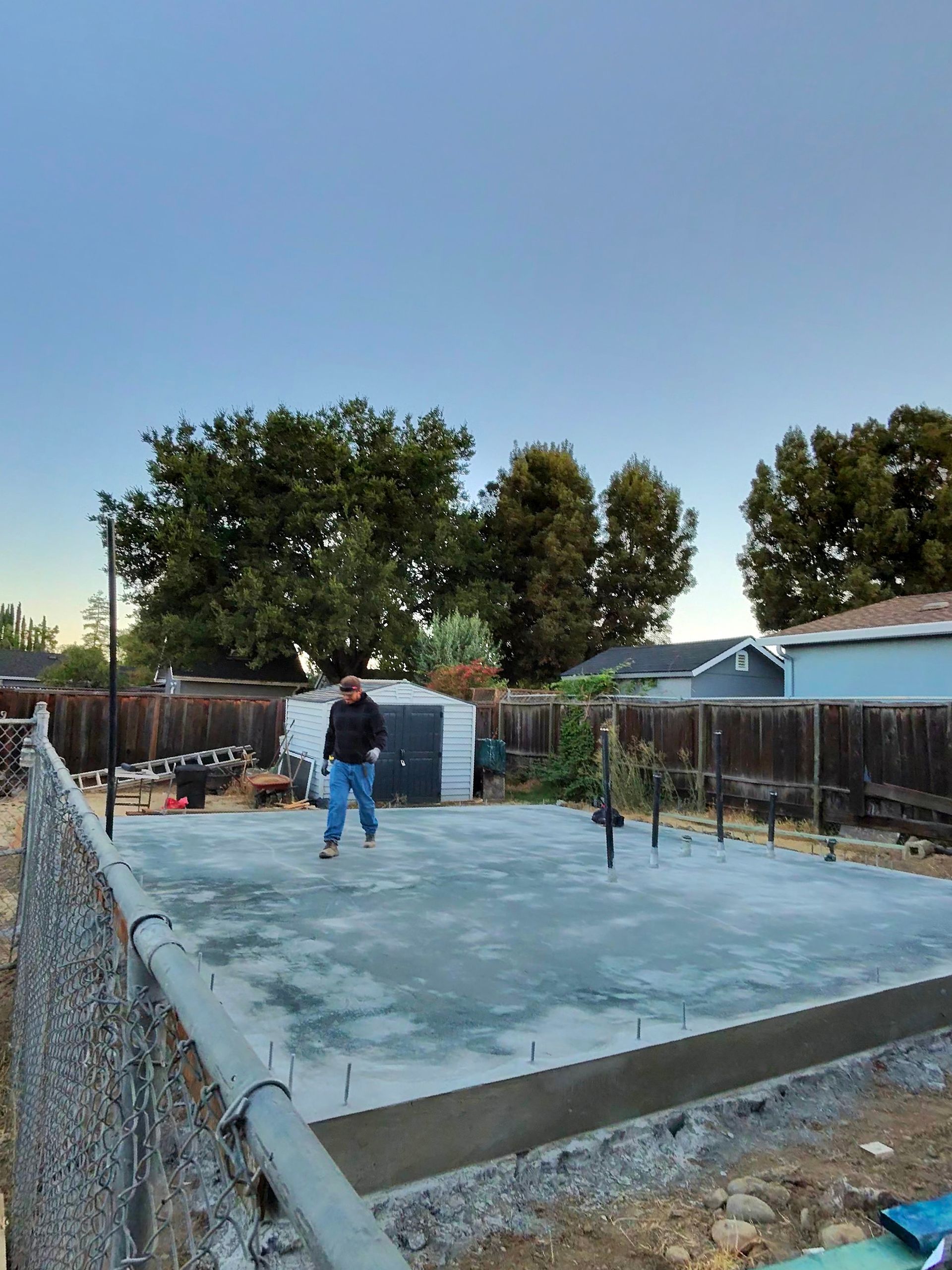 Person standing on a new concrete slab in a fenced backyard at dusk
