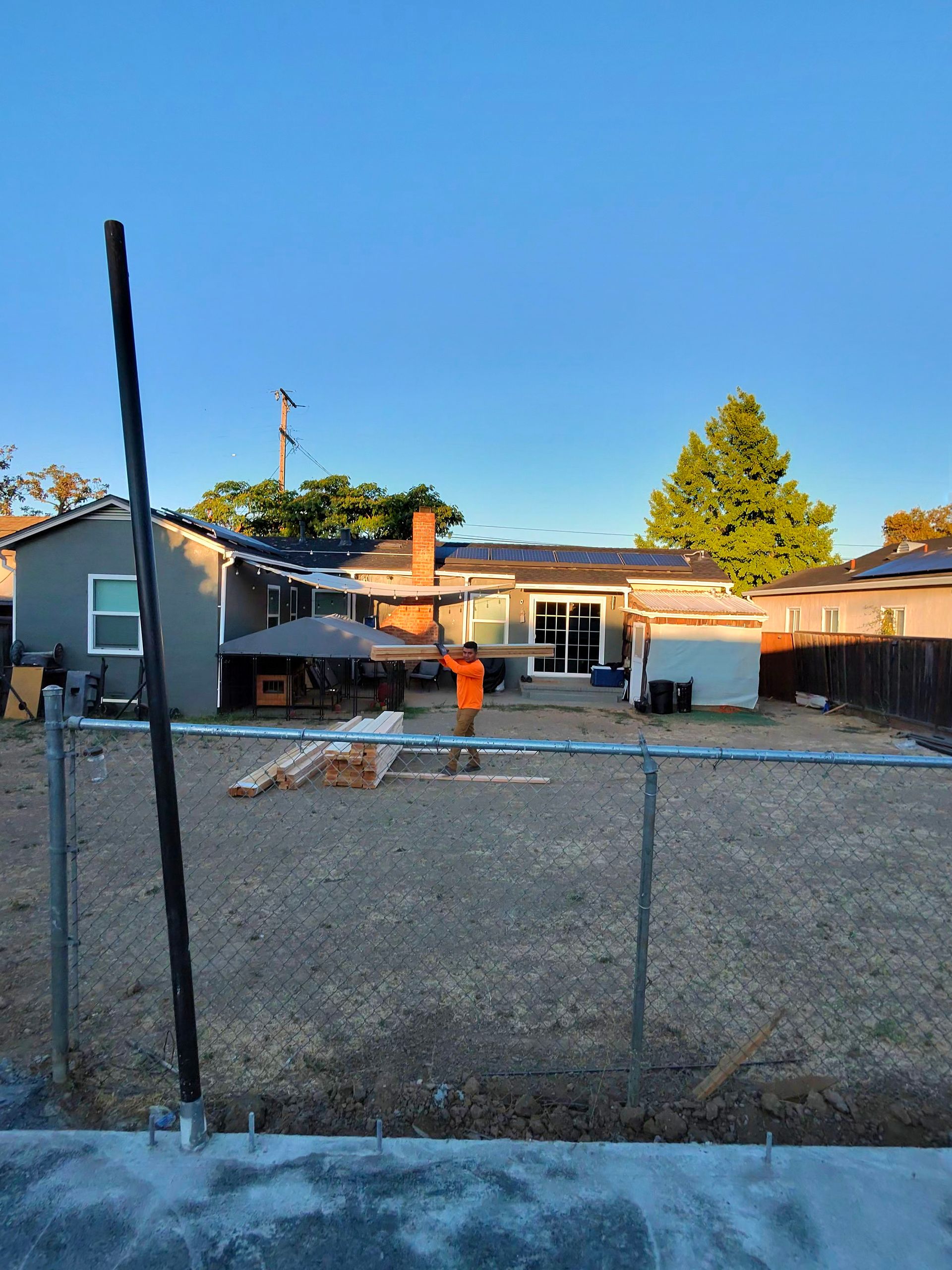 Chain-link fence in front of single-story houses at sunset, with a tall pole in the foreground.