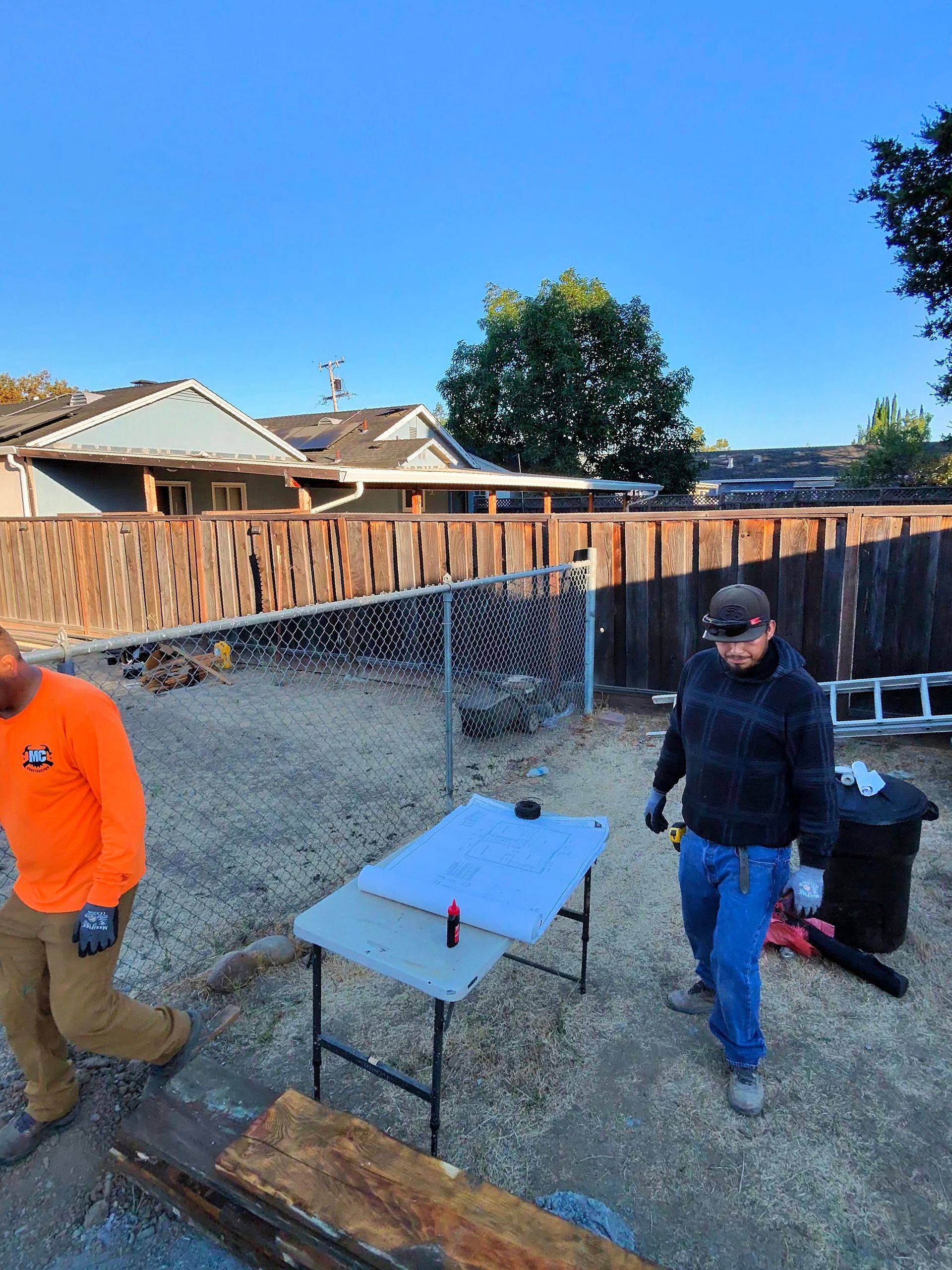 Backyard worksite with two people, a table, tools, gravel, and a wooden fence under a clear sky