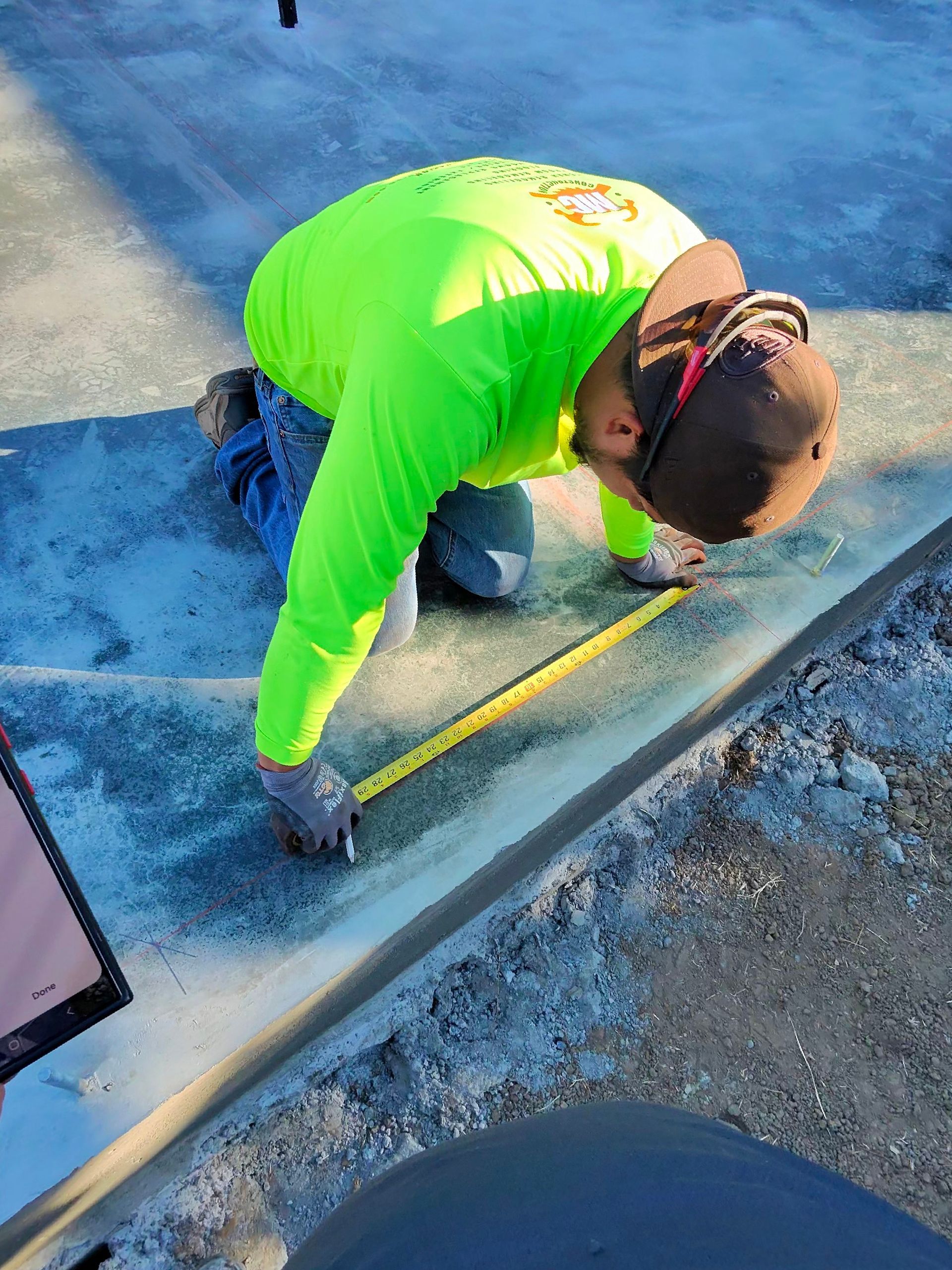Worker in neon green shirt kneeling and measuring along a concrete edge outdoors with a tape measure