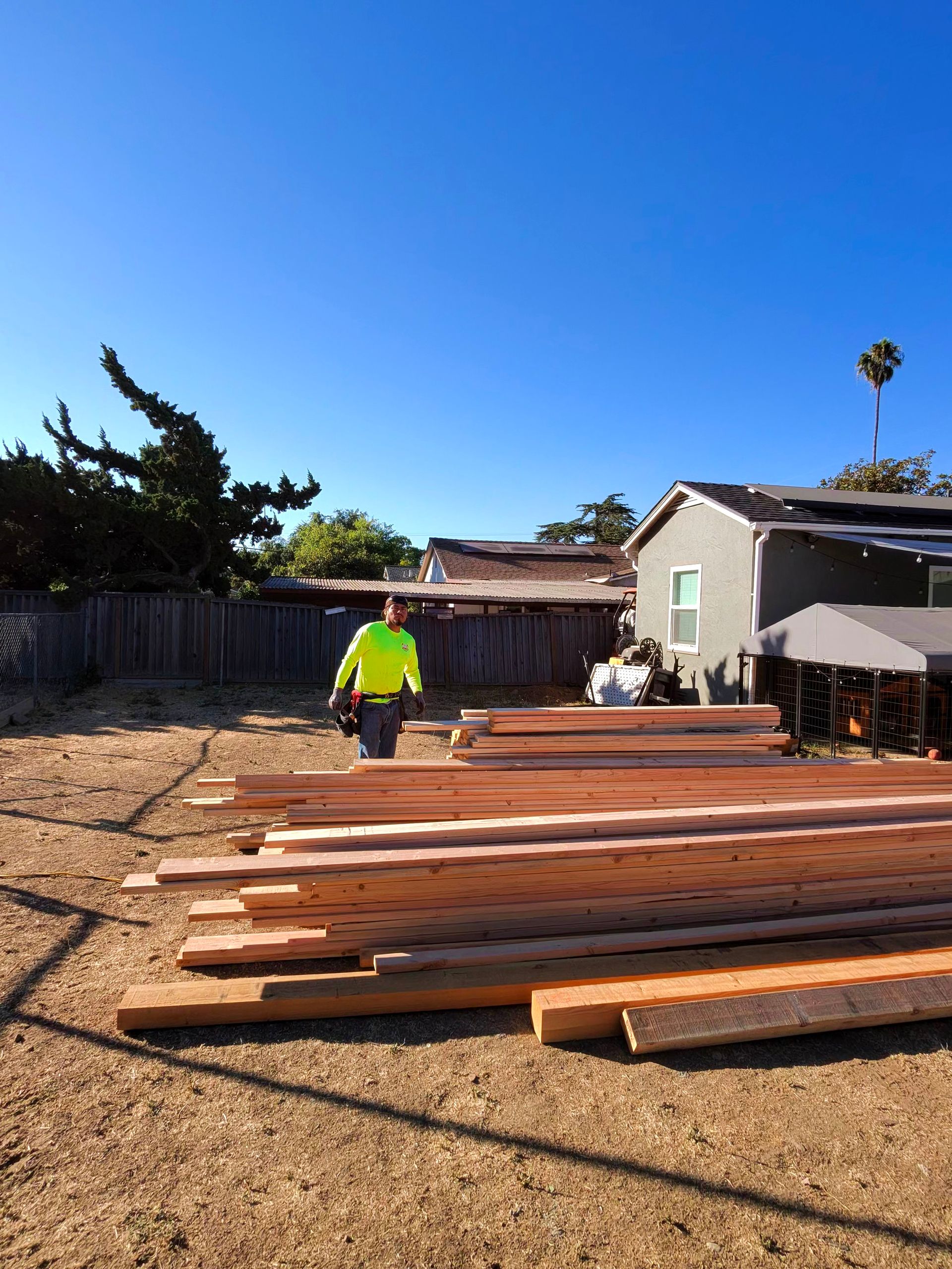Wooden beams stacked in a backyard while a worker in a neon shirt stands nearby under a clear blue sky