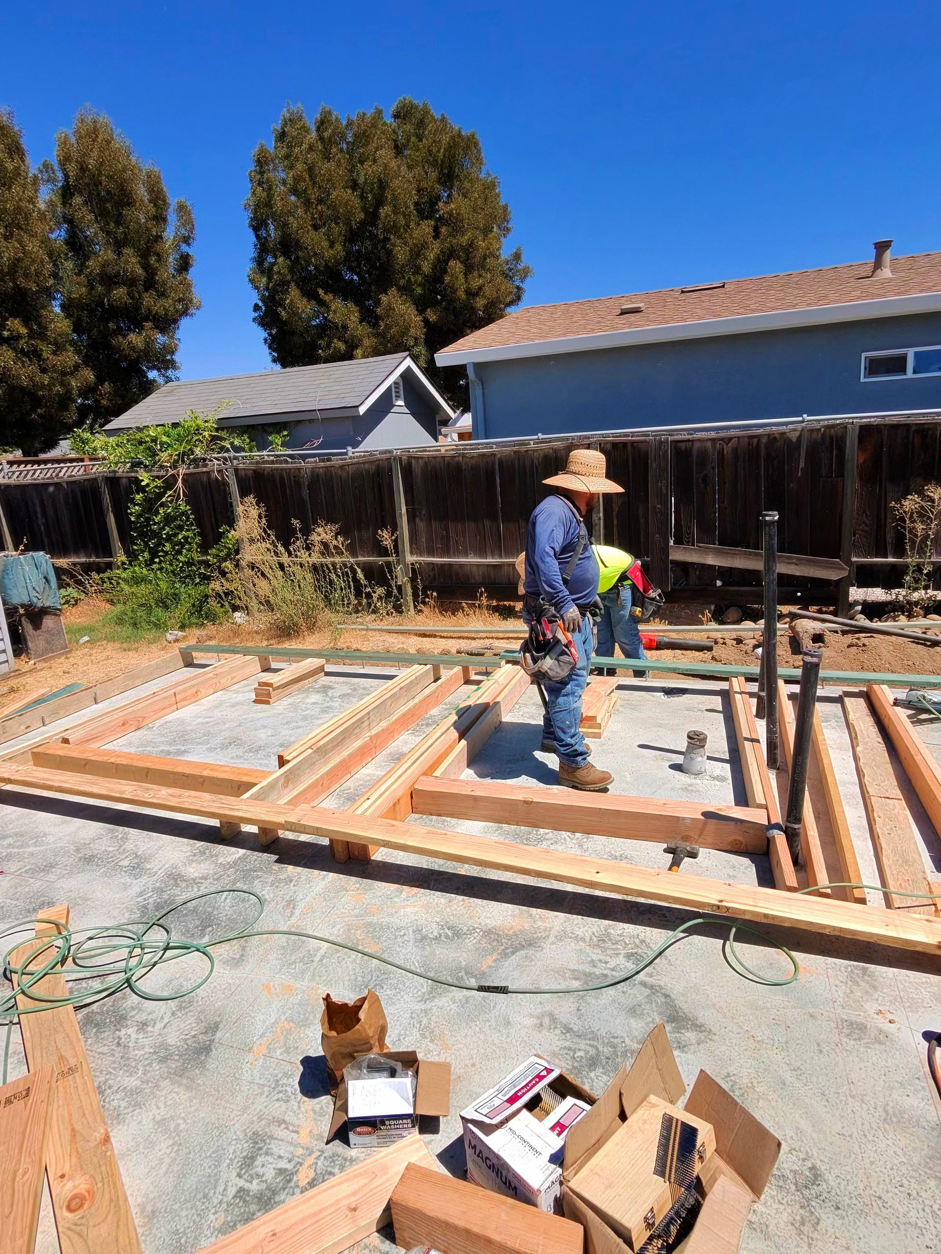 Workers building a house foundation with wooden framing in a sunny backyard, tools and materials scattered nearby