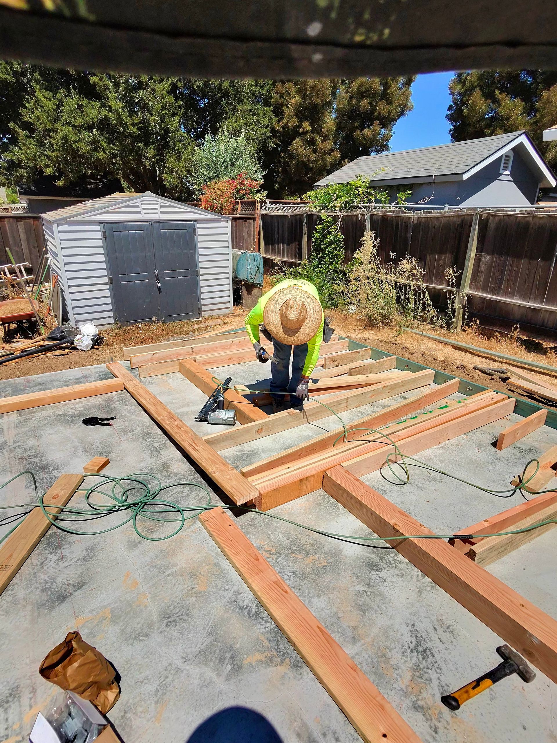 Worker in a straw hat measuring lumber on a backyard deck frame under construction.