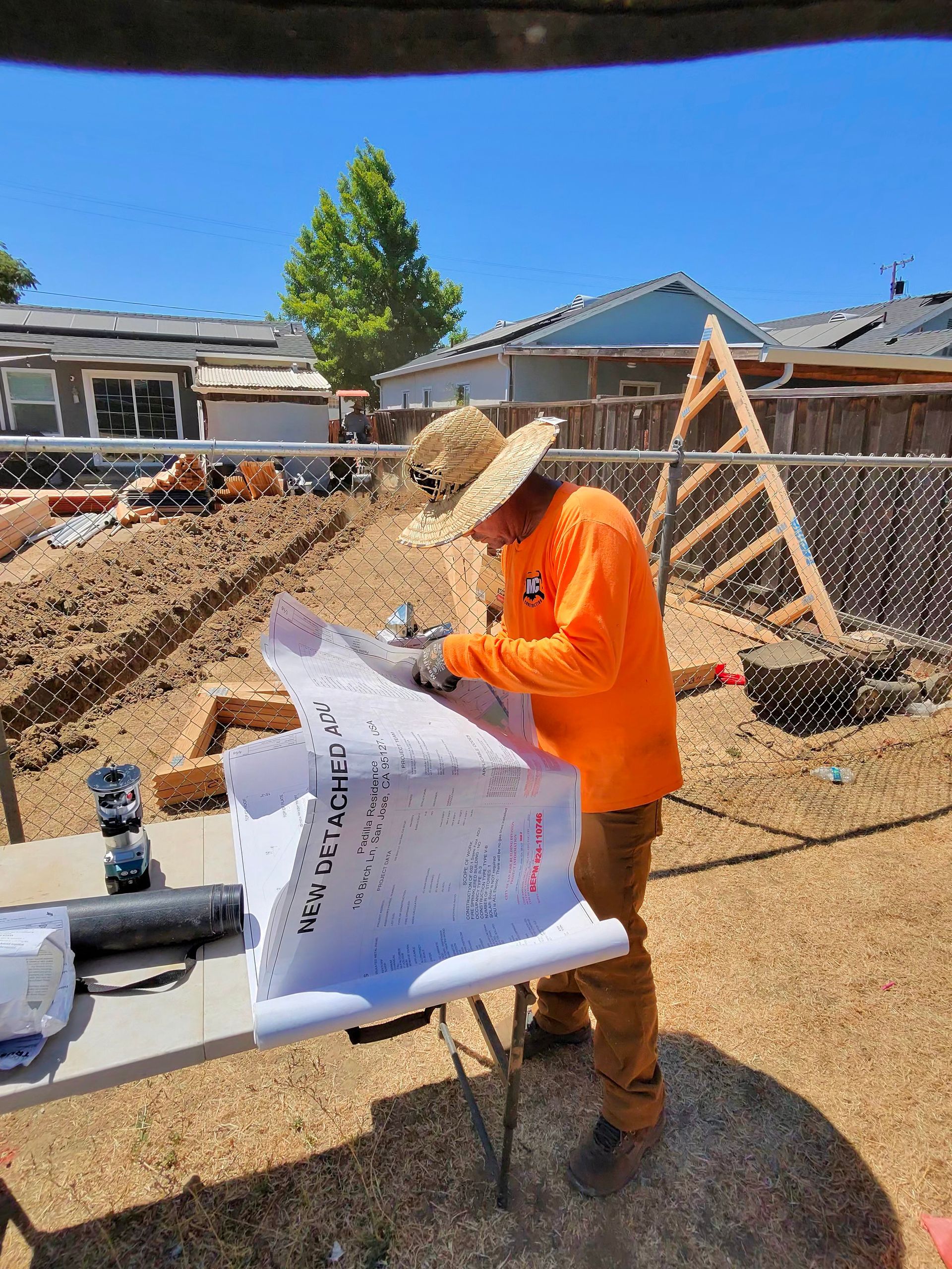 Construction worker in orange shirt reading plans on a job site under bright blue sky