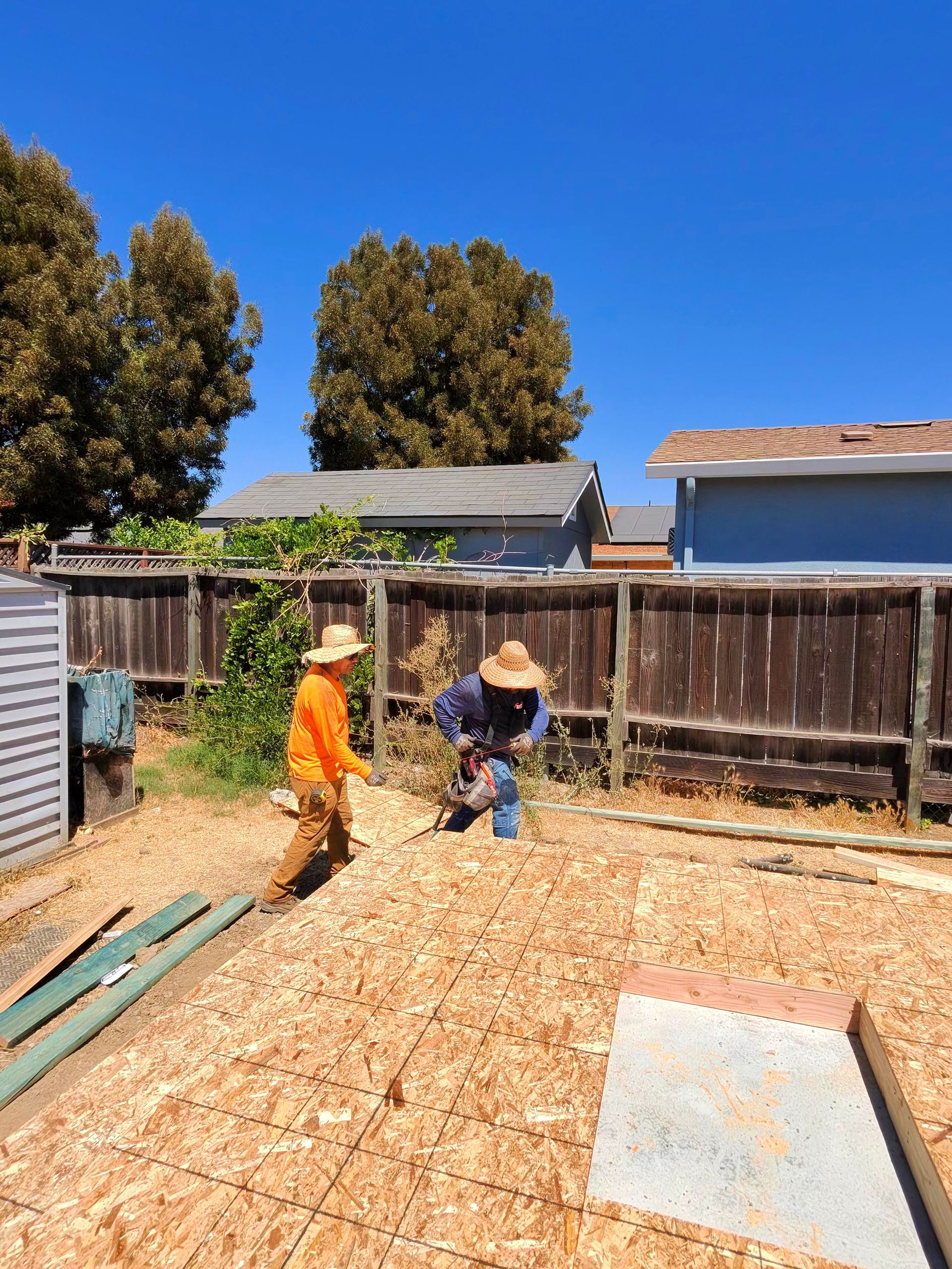 Two workers in hard hats clearing wood chips in a backyard under a clear blue sky