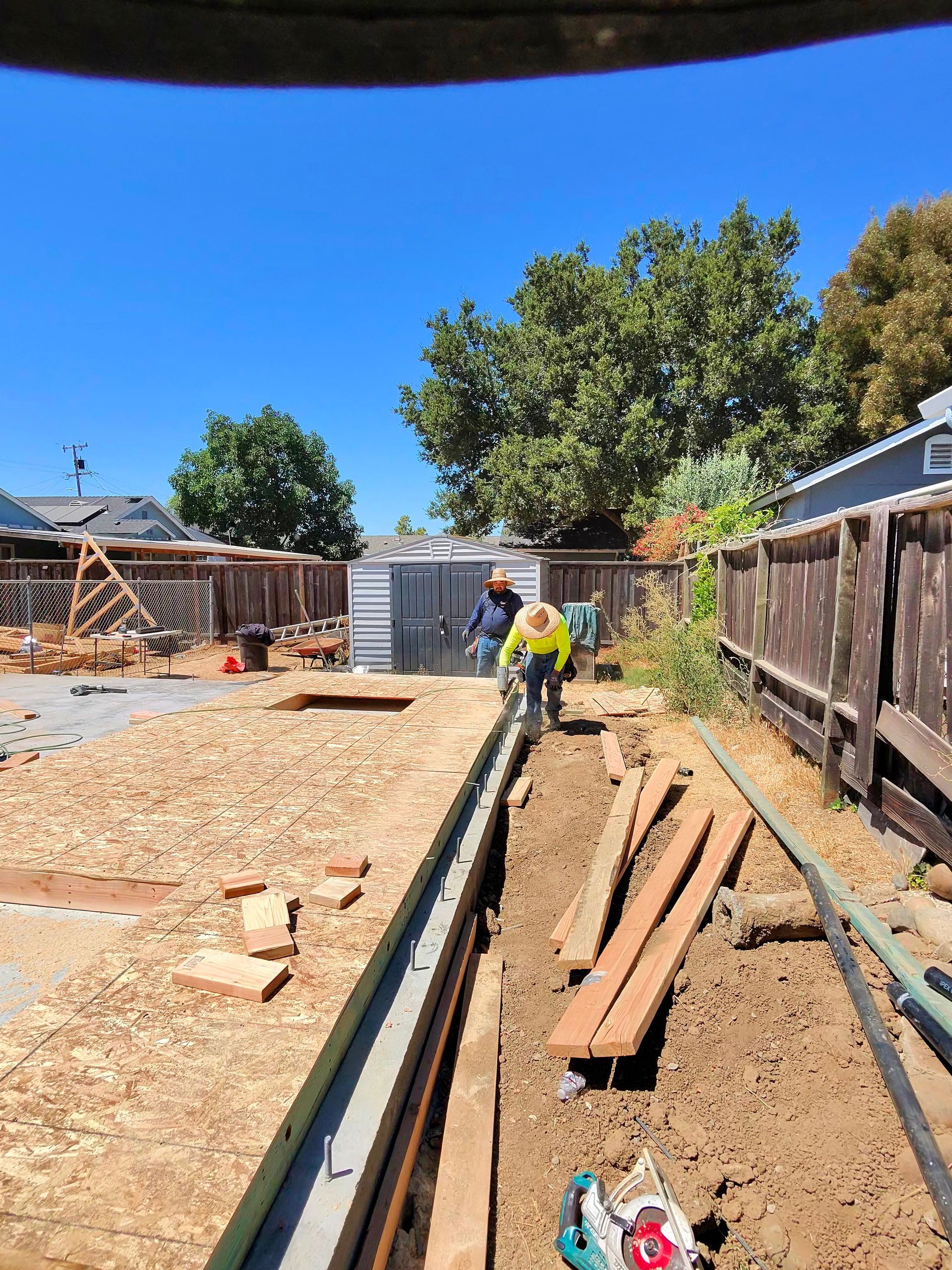 Construction workers installing a long concrete curb along a dirt path in a backyard under a clear blue sky
