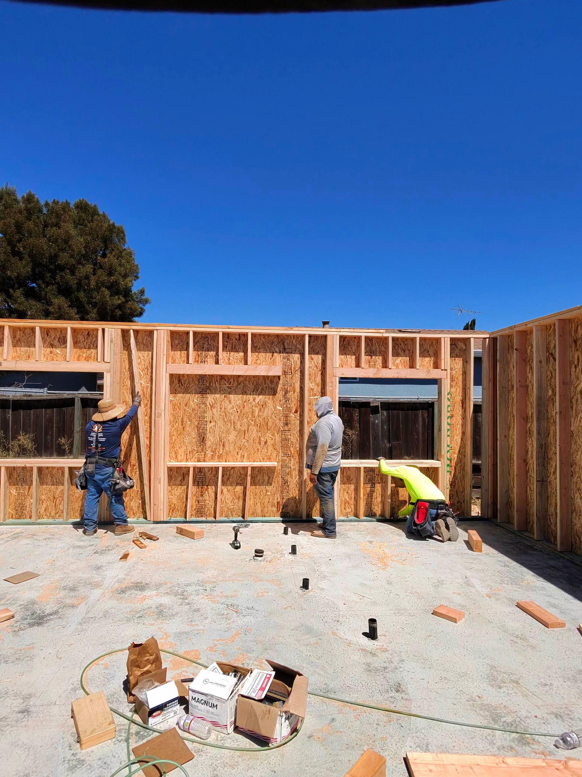 Construction workers framing a house on a gravel lot under a clear blue sky