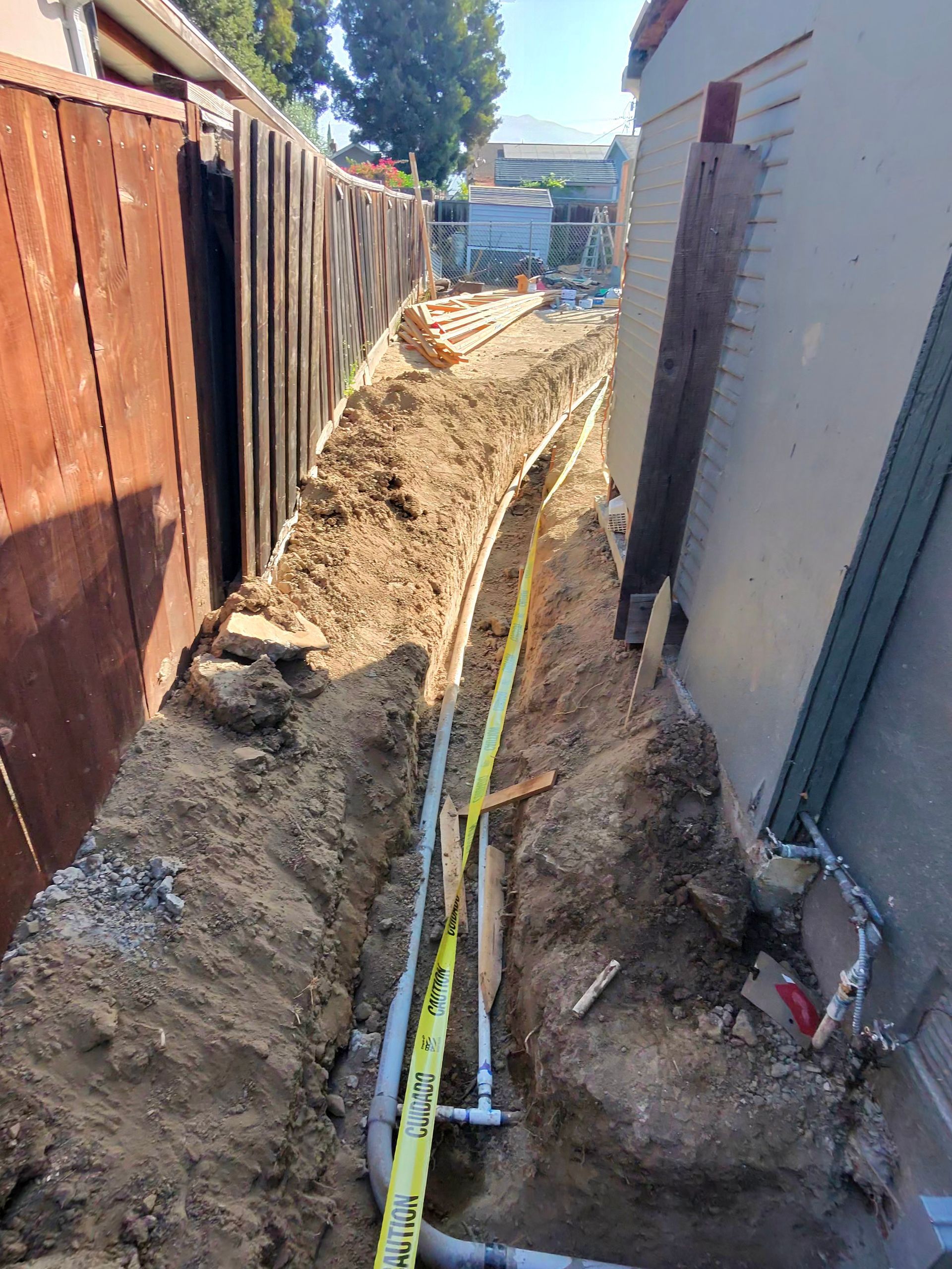Narrow backyard trench beside a house and fence, with exposed dirt and utility lines under construction