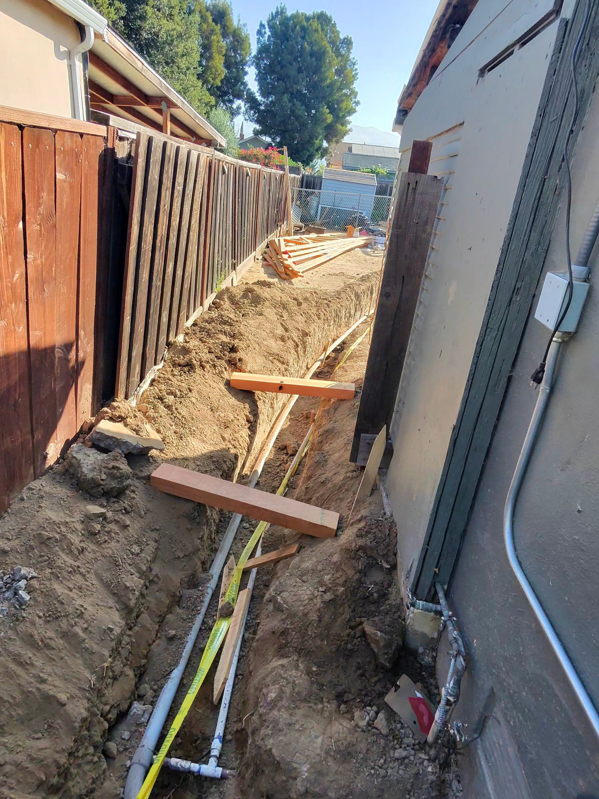 Narrow trench between houses with exposed pipes, wooden braces, and dirt from an excavation site