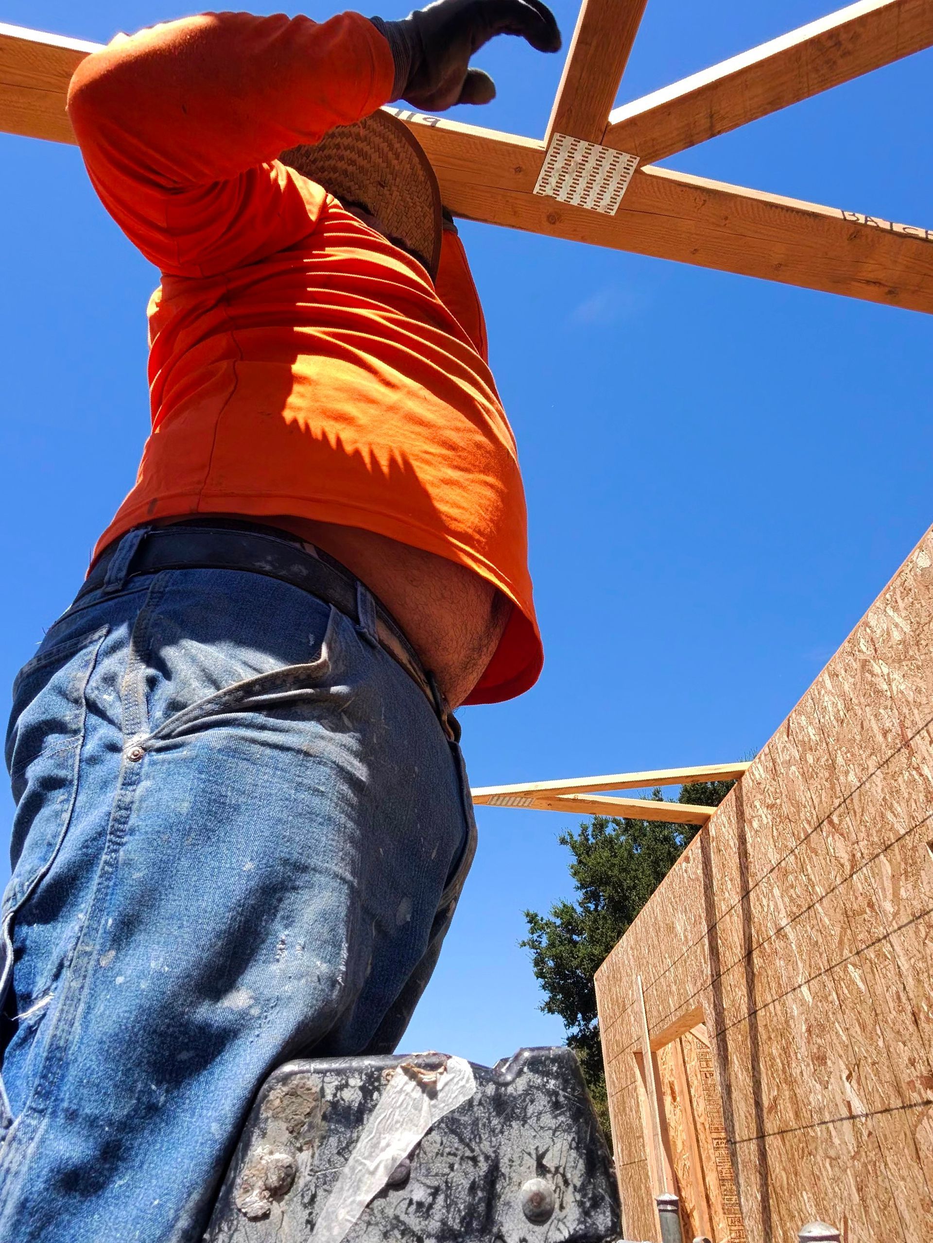 Person in orange shirt and jeans working on a wooden construction frame against a blue sky.